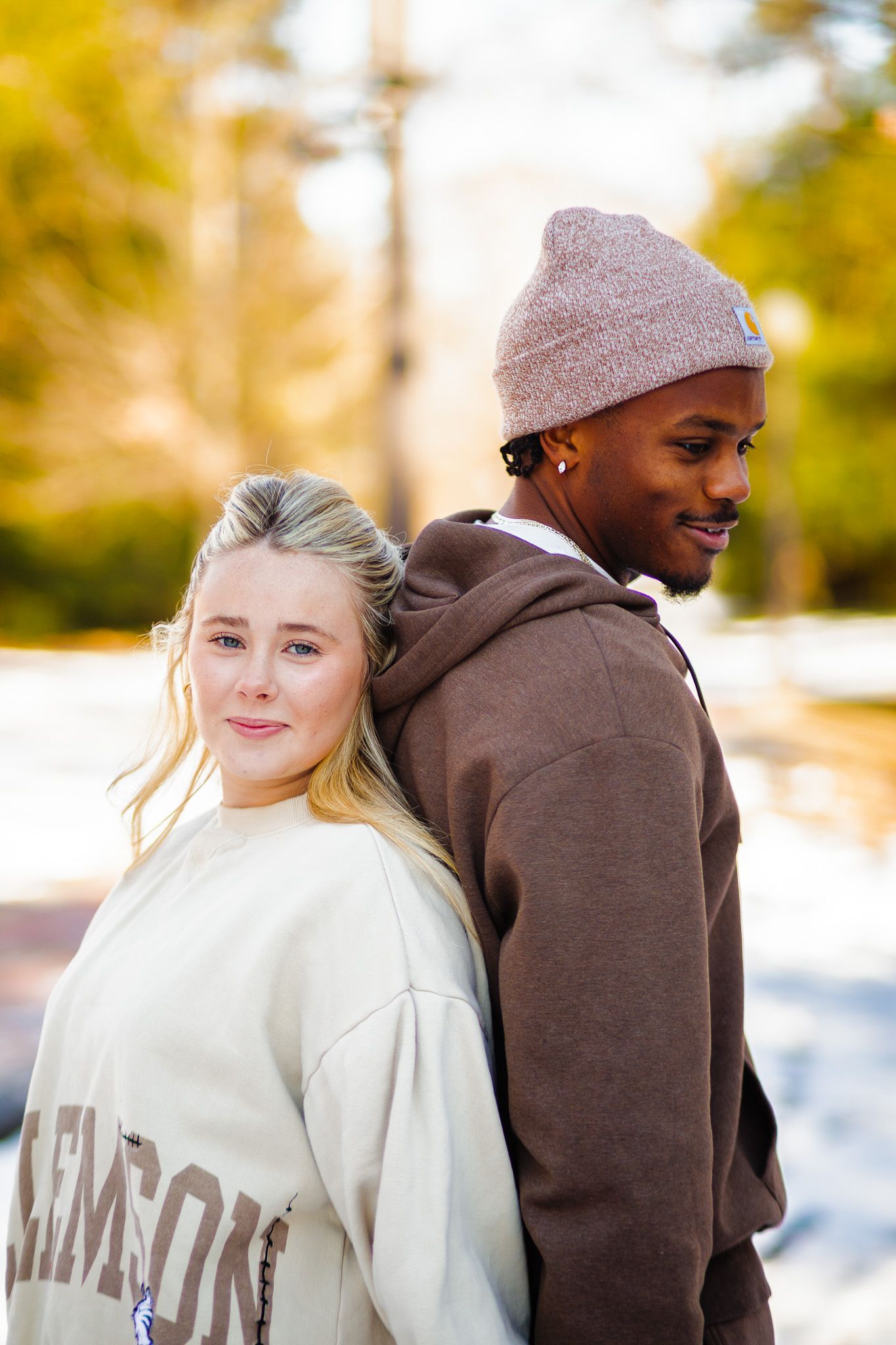 Two young adults in casual hoodies pose back-to-back outdoors, smiling, with autumn trees in the background.