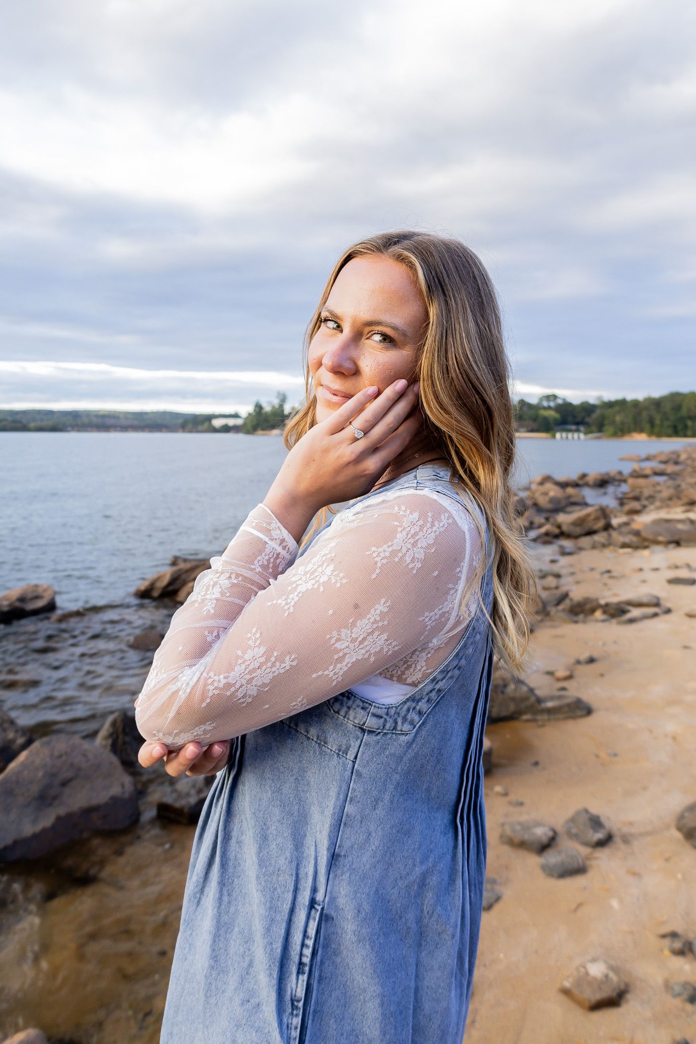 Young woman in lace top and denim overalls poses on rocky beach shoreline at golden hour.