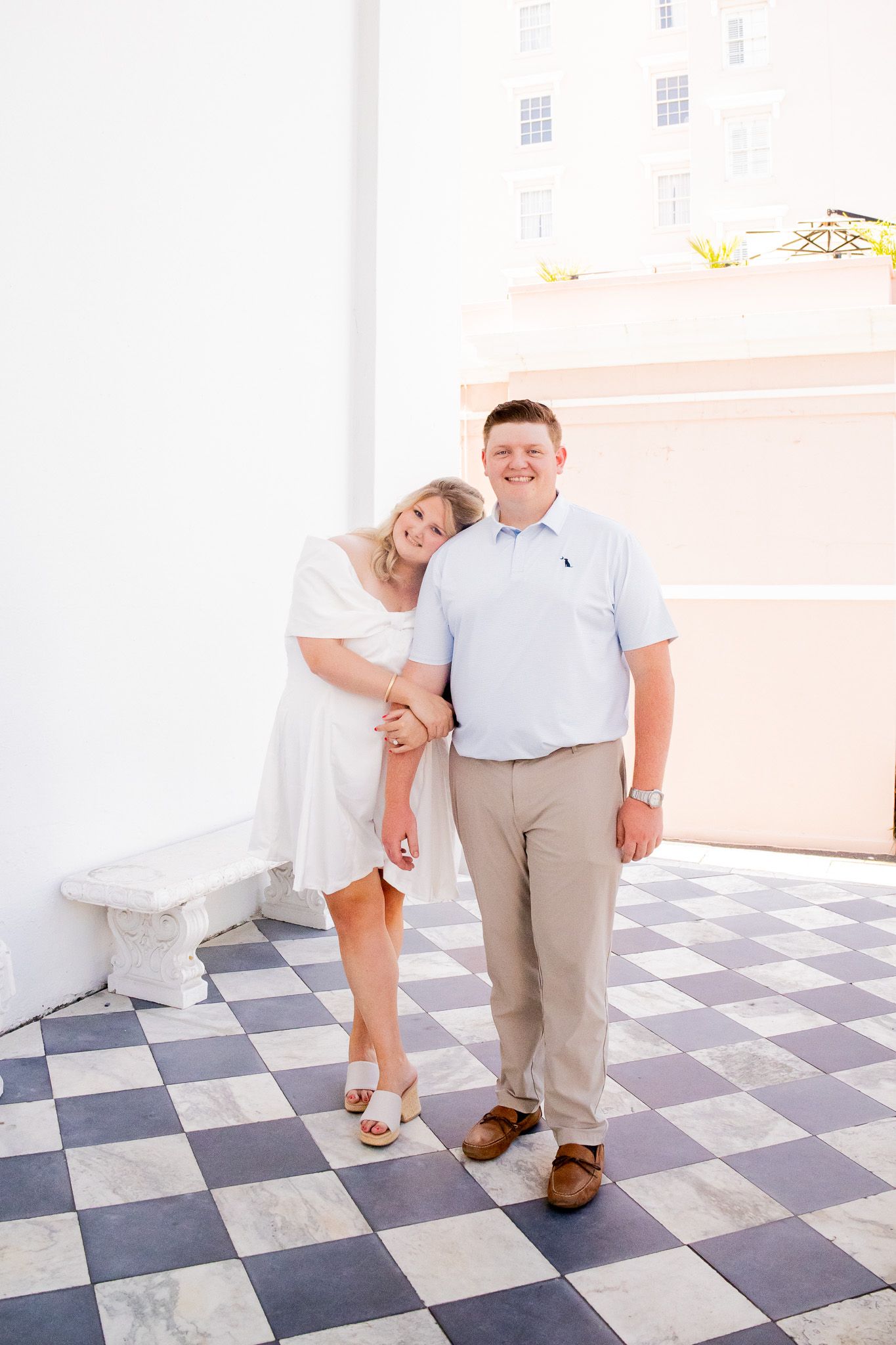 Couple posing on checkered floor, woman in white dress leaning on man in light blue polo and khakis.