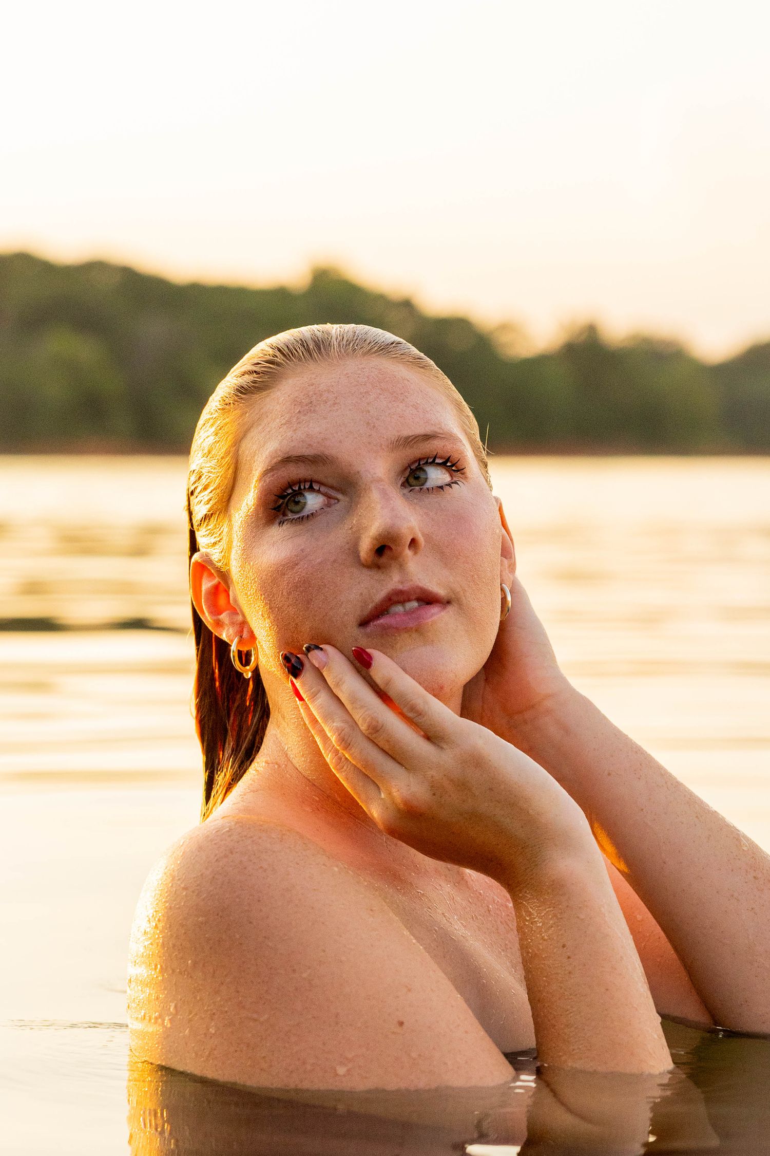 Young woman with wet hair poses in a lake at golden hour sunset, hands framing her face.