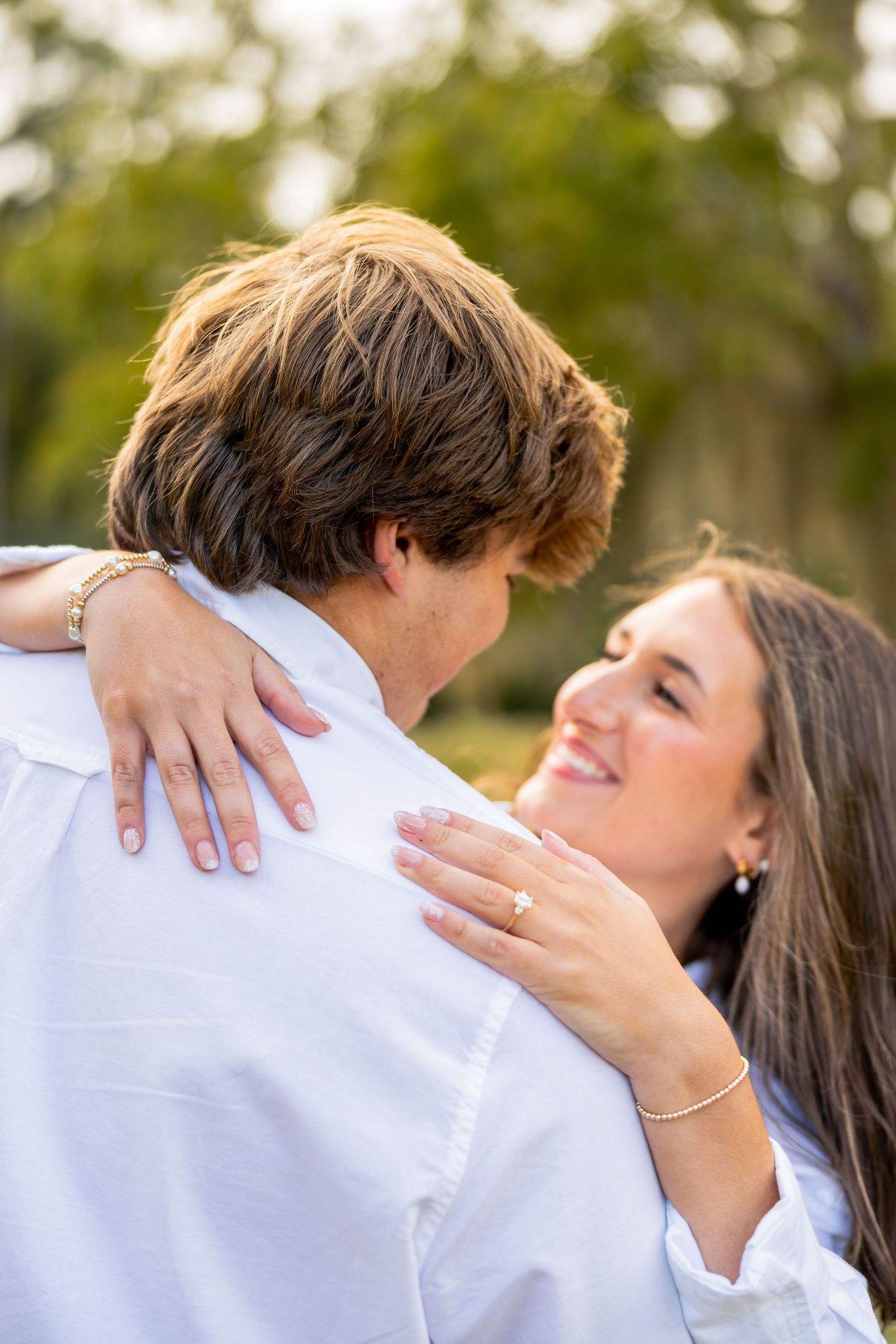 Smiling woman with a diamond ring embraces a man in a white shirt outdoors, a joyful engagement moment against a soft green background.