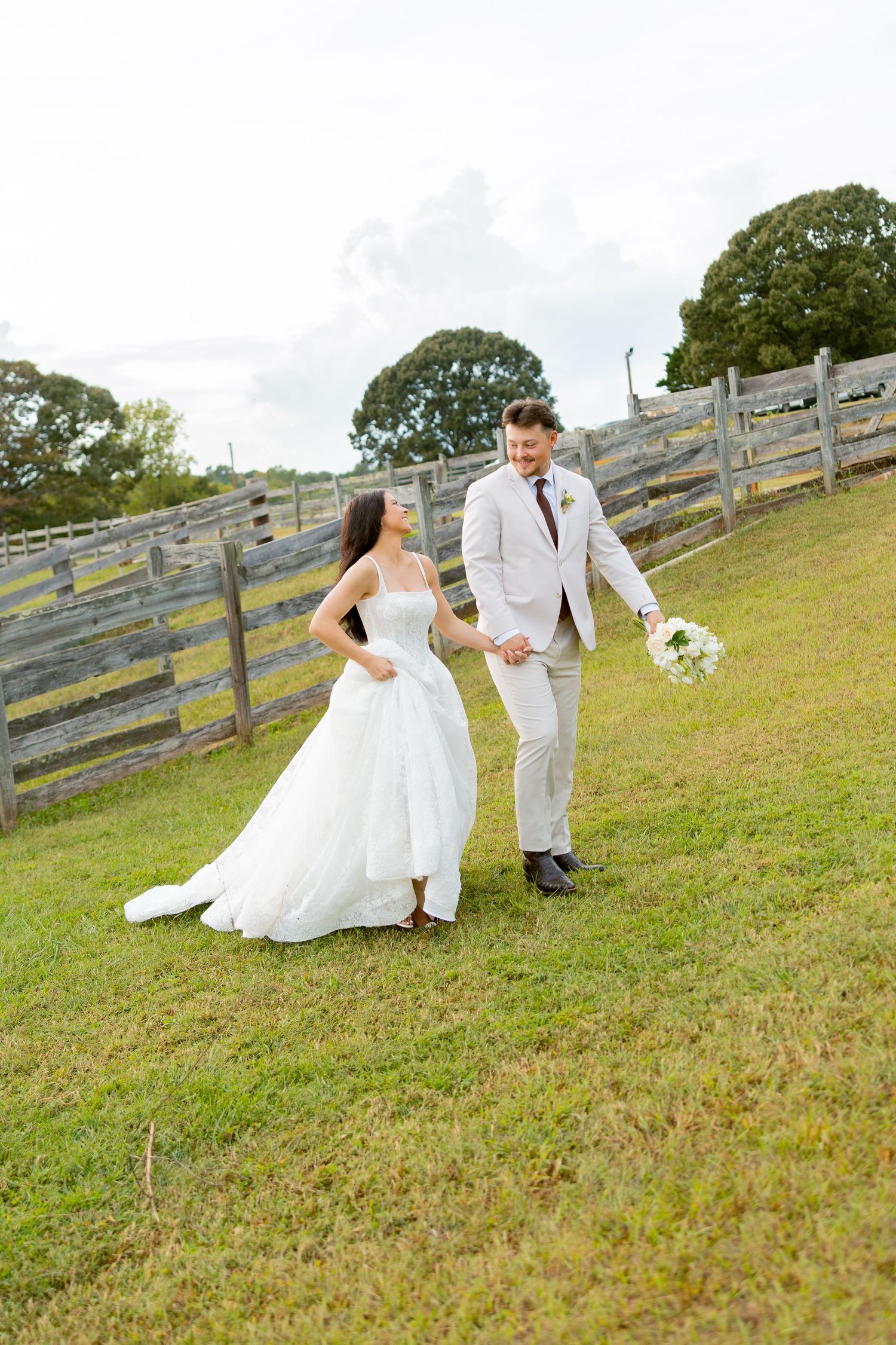 A bride and groom walk hand-in-hand through a grassy field beside a rustic wooden fence on their wedding day.