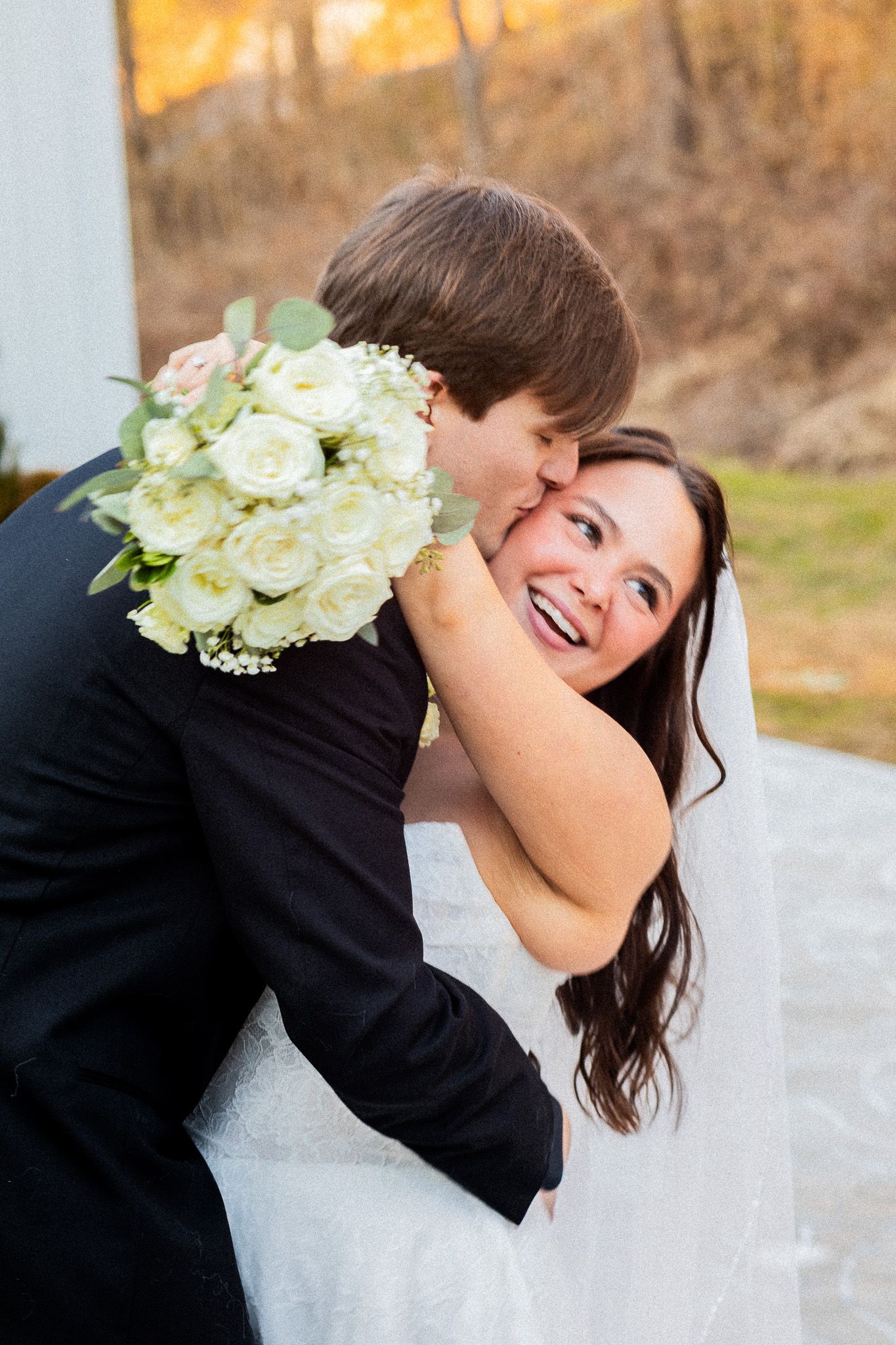 Groom in black suit kissing bride on cheek as she smiles holding white rose bouquet on wedding day.