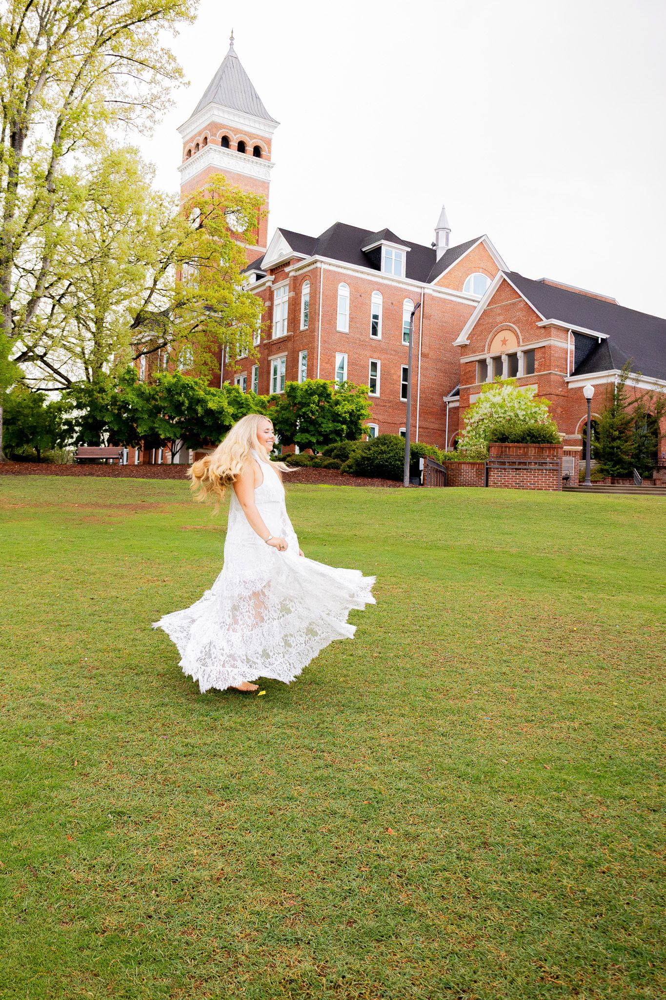 Bride in white lace gown twirling on green lawn before a red brick building with a clock tower.