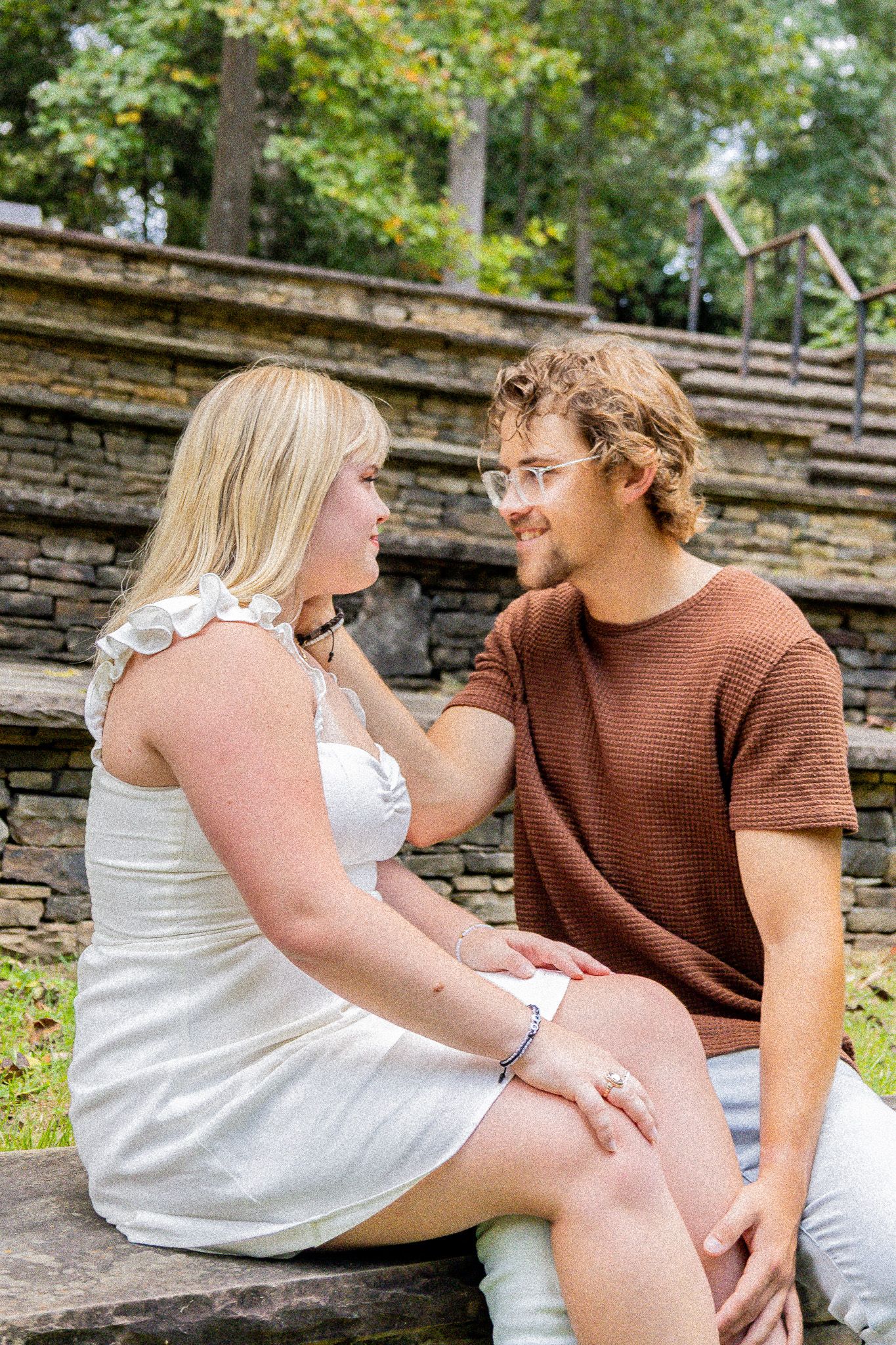 A young couple sits together on stone steps outdoors, smiling at each other in a romantic moment.