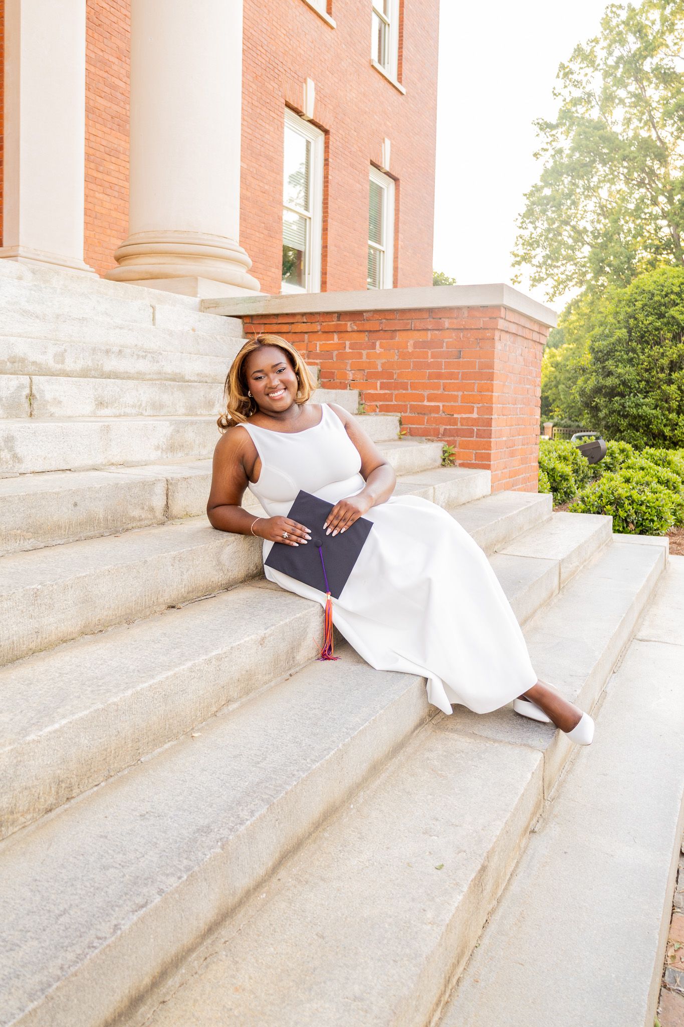 Smiling Black woman in white dress sits on stone steps holding graduation cap outside brick building.