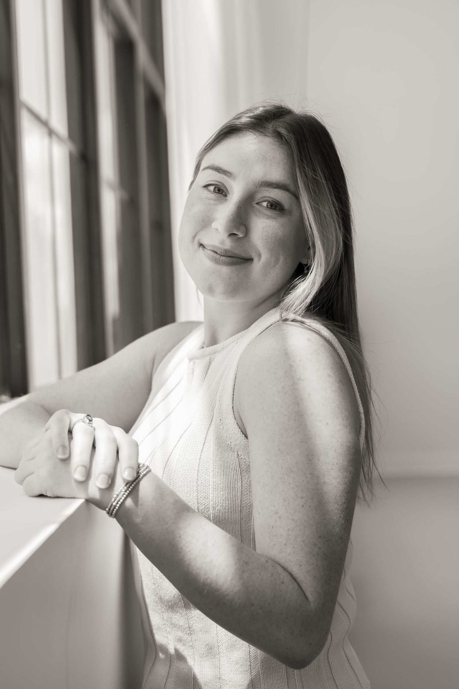 Black and white portrait of a smiling young woman leaning against a windowsill in a sleeveless top.