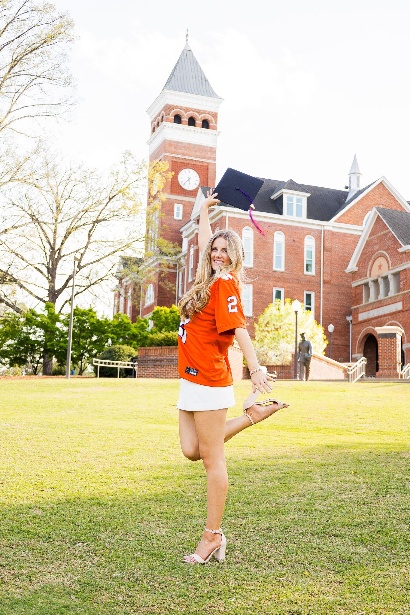 Smiling blonde graduate in orange jersey holding cap while posing on campus lawn near brick clock tower.