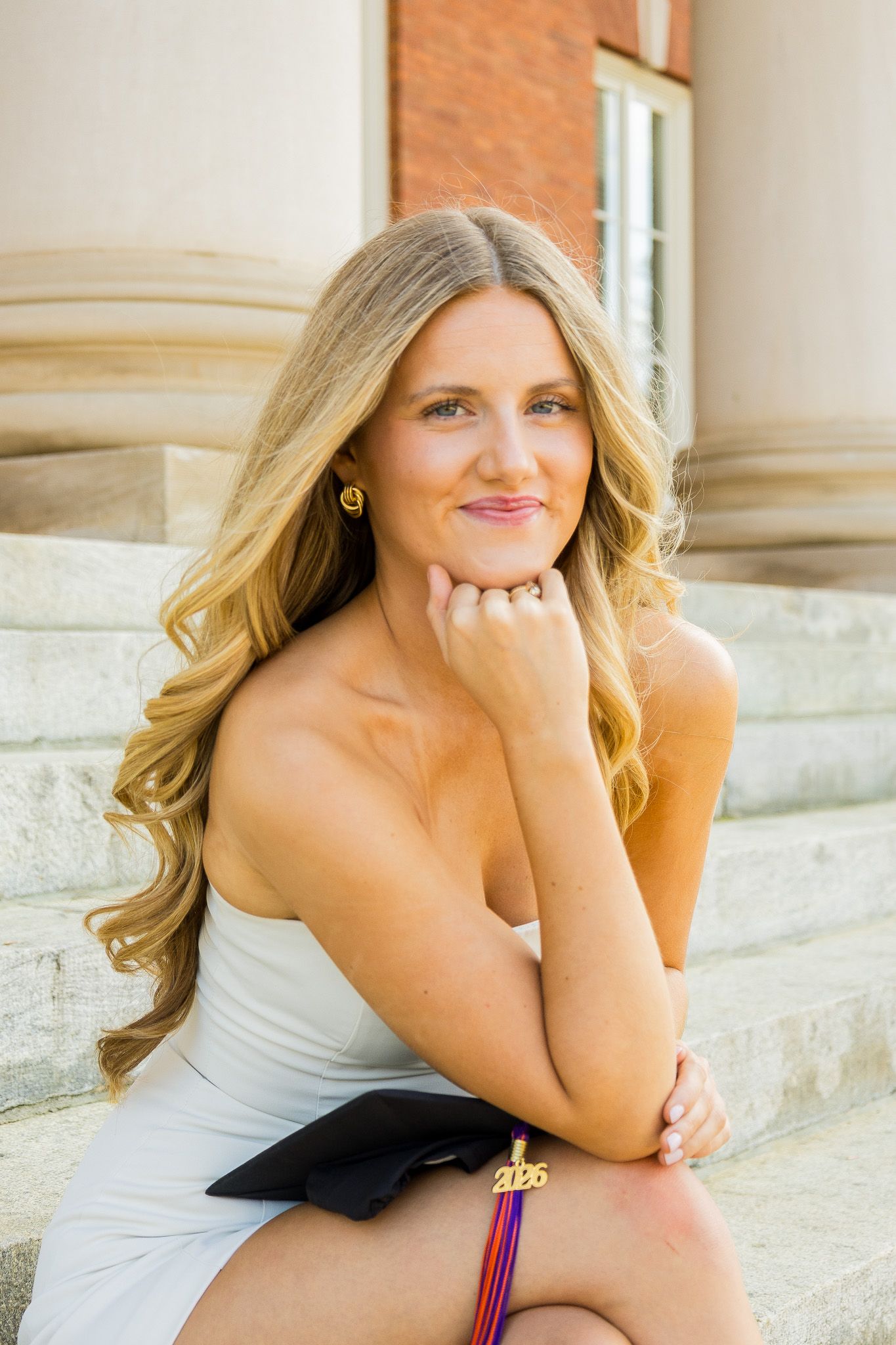 Young blonde woman in white dress holds graduation cap on stone steps, smiling confidently at her 2025 graduation photo.