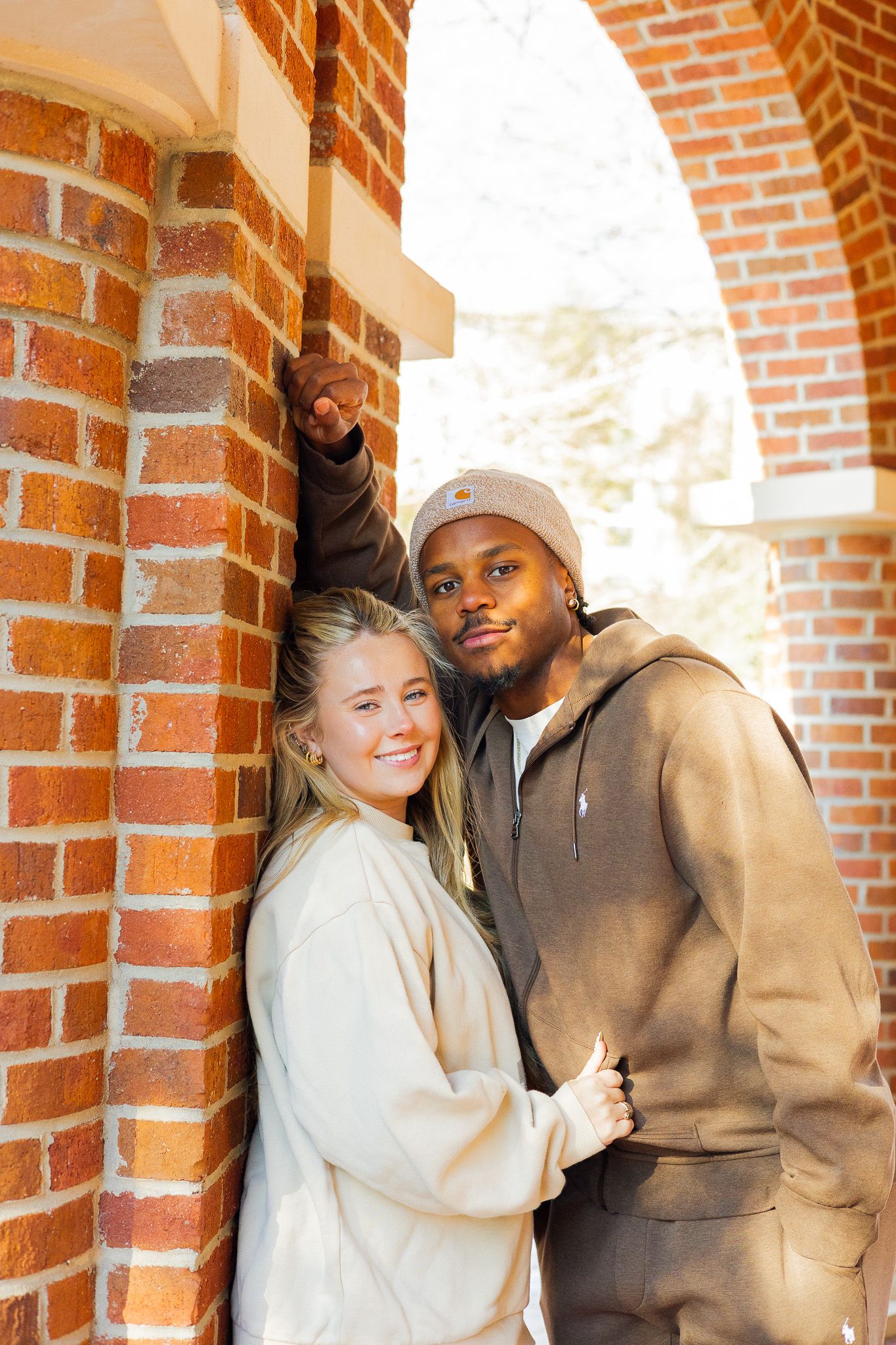 A smiling interracial couple poses together against a red brick archway outdoors.