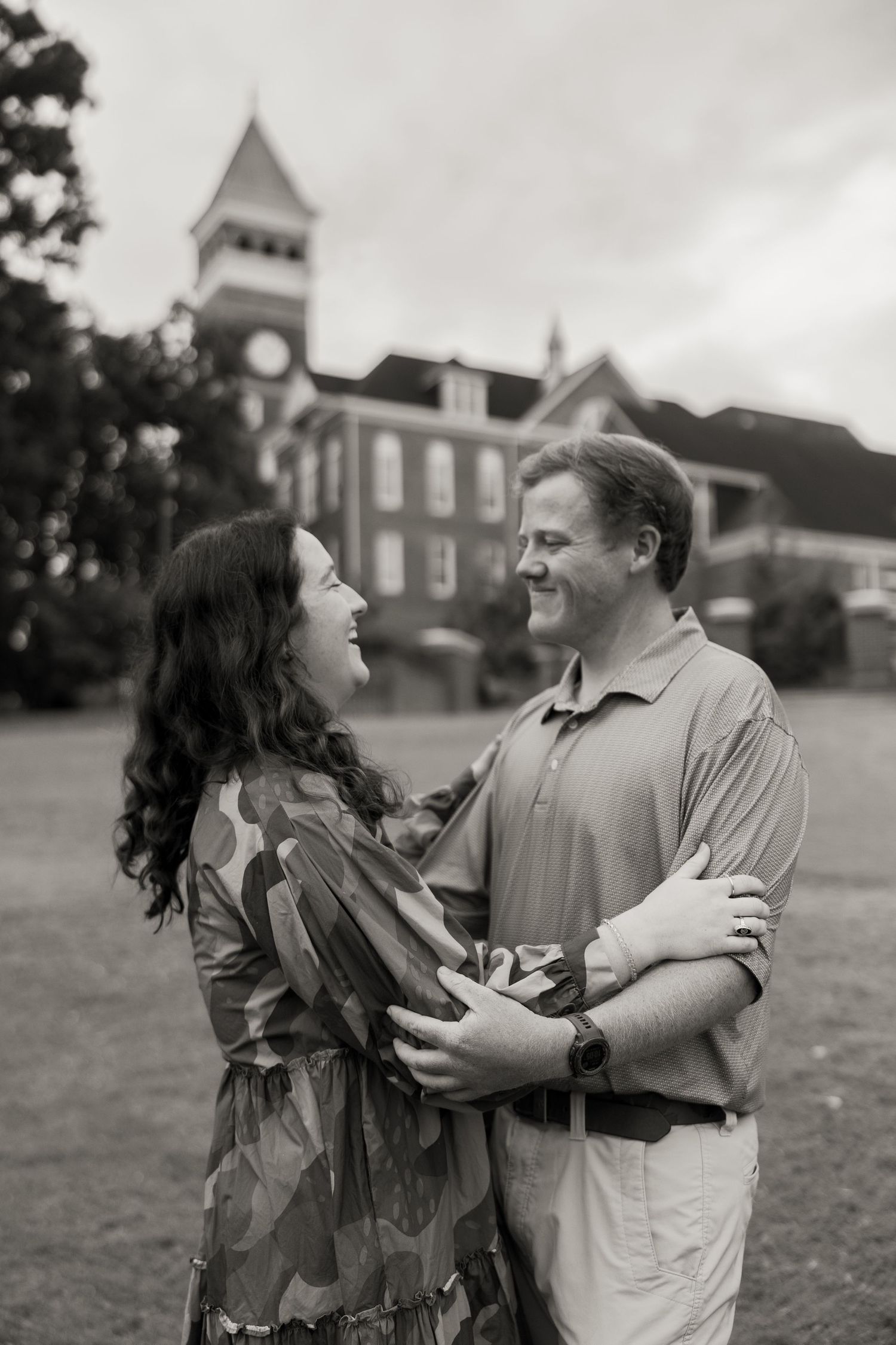Black and white photo of a couple embracing and smiling at each other in front of a historic brick building.
