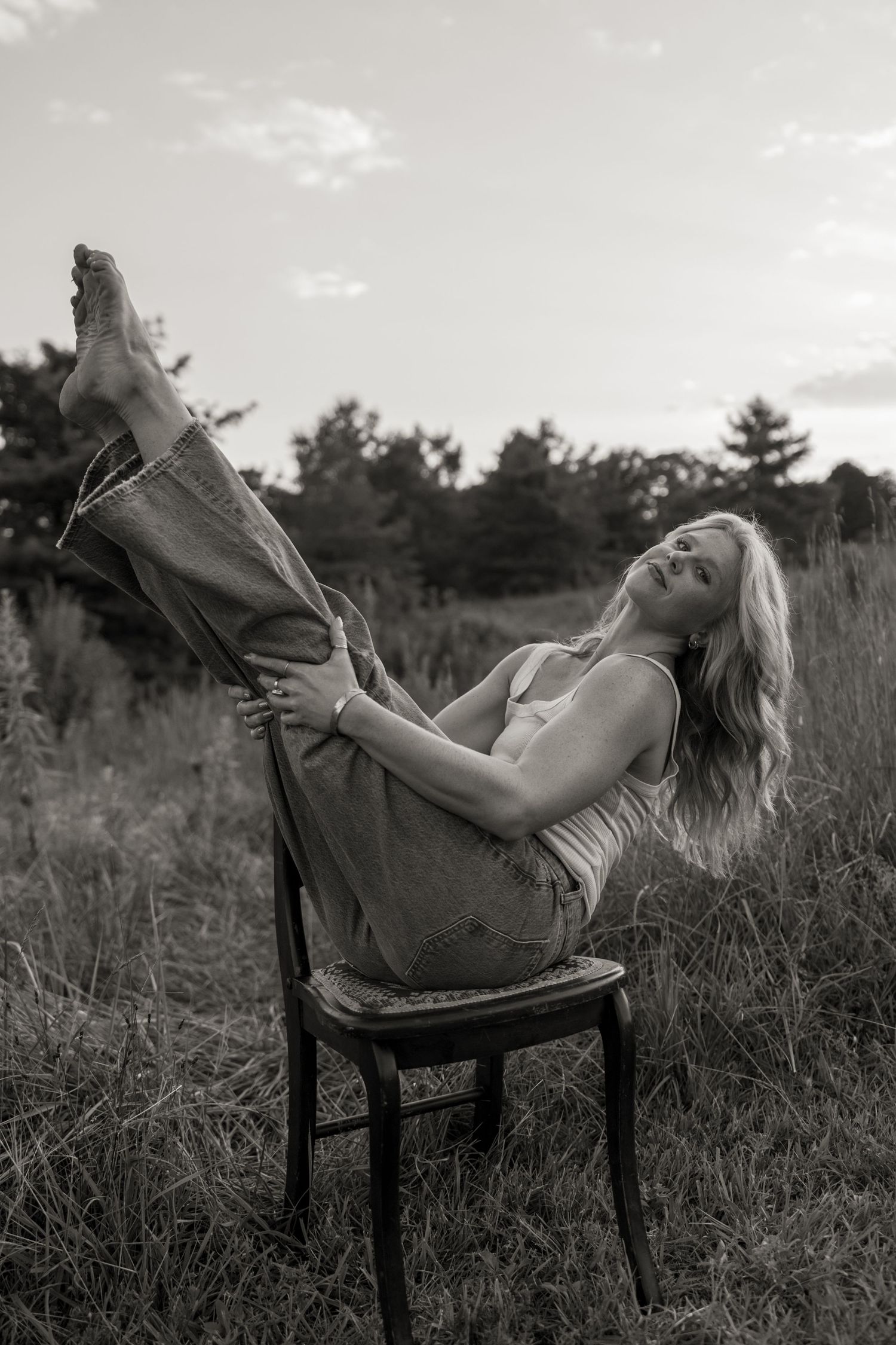 Black and white photo of a woman sitting on a wooden chair in a field, legs raised, wearing jeans and a tank top.
