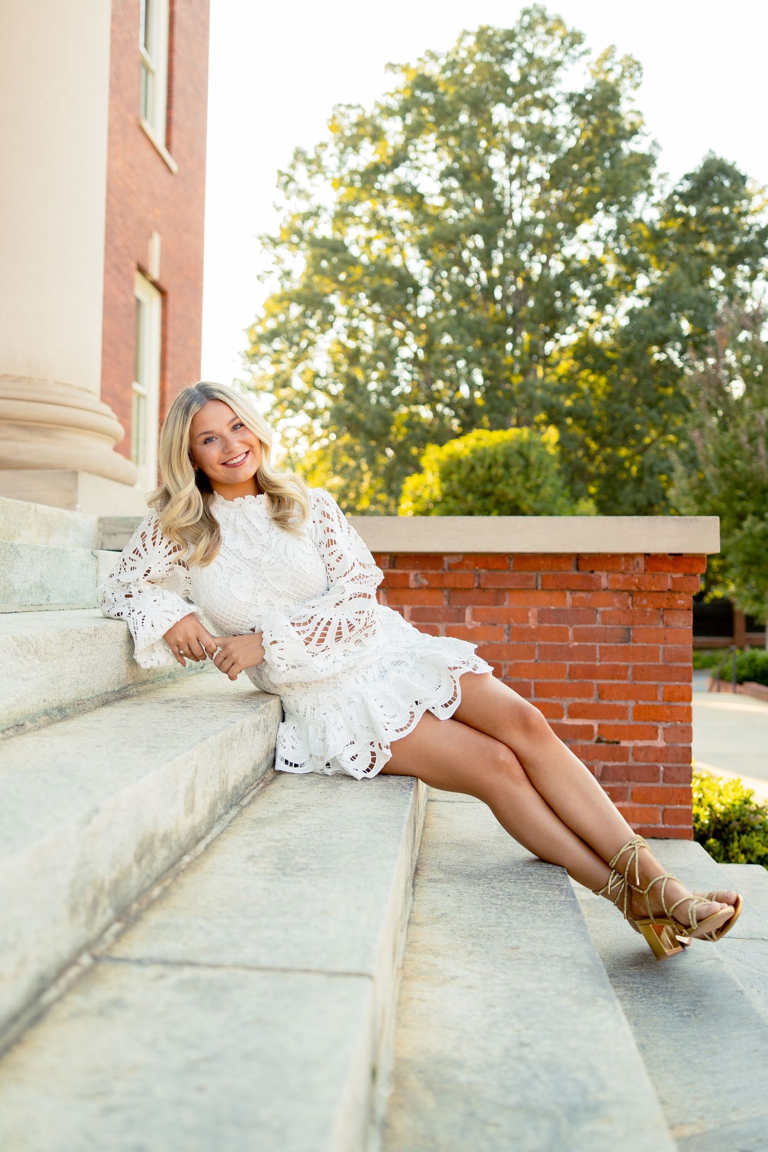 Young woman in white lace dress and heels sitting on stone steps, smiling outdoors near brick building.