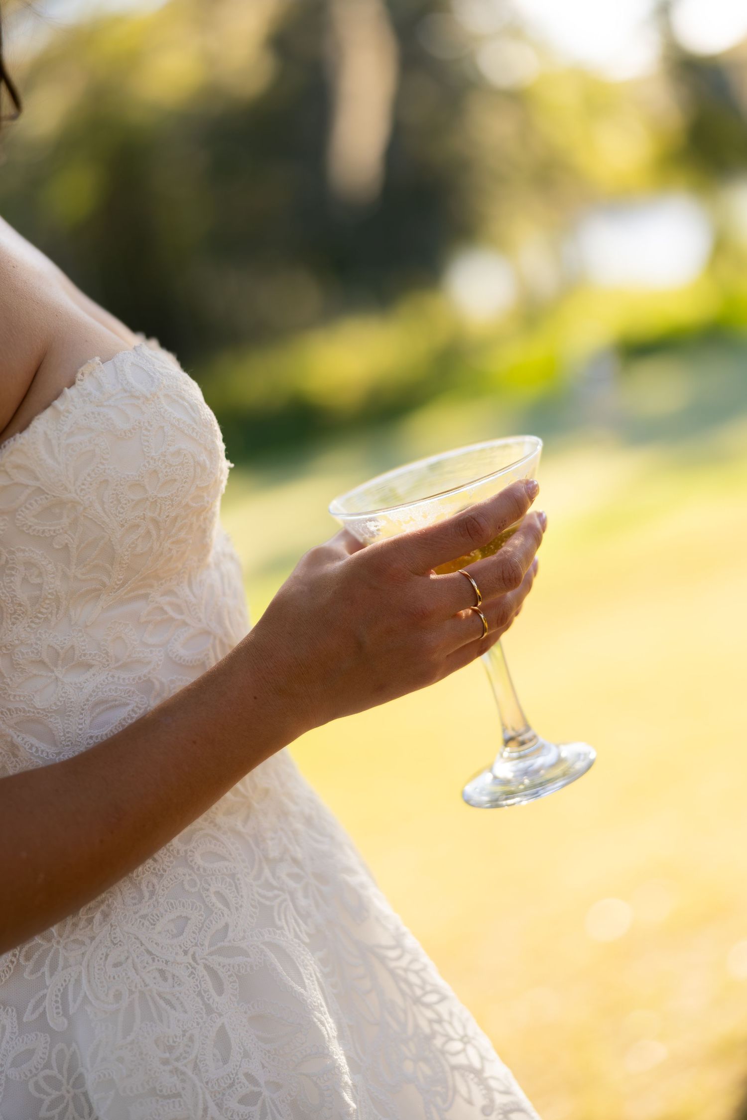 Bride in white lace dress holding a champagne glass outdoors in golden sunlight, with a gold wedding ring visible.