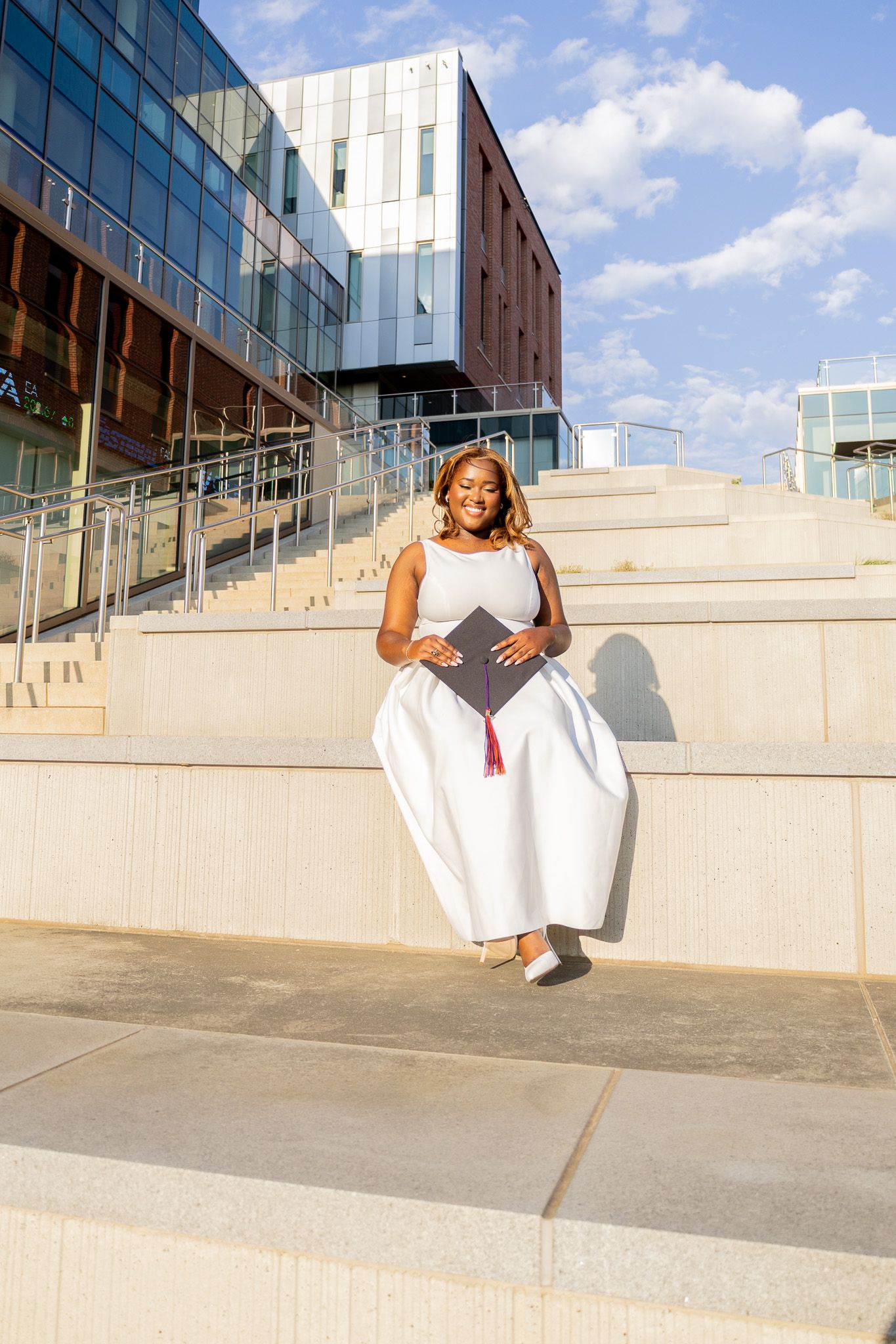 Young Black woman in white dress holds graduation cap on university steps, celebrating academic achievement outdoors.