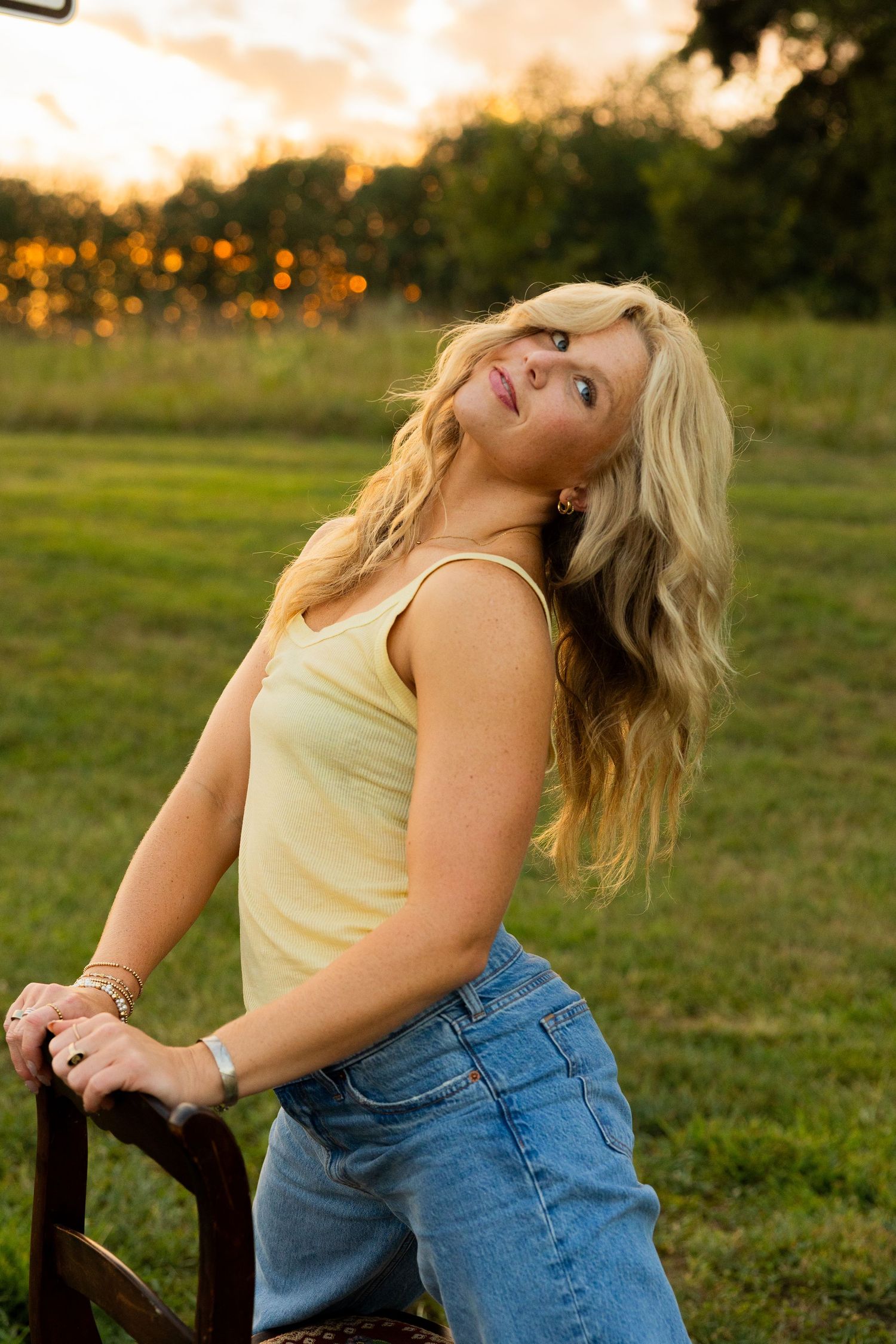 Blonde woman in yellow tank top and jeans posing outdoors at sunset in a green field.