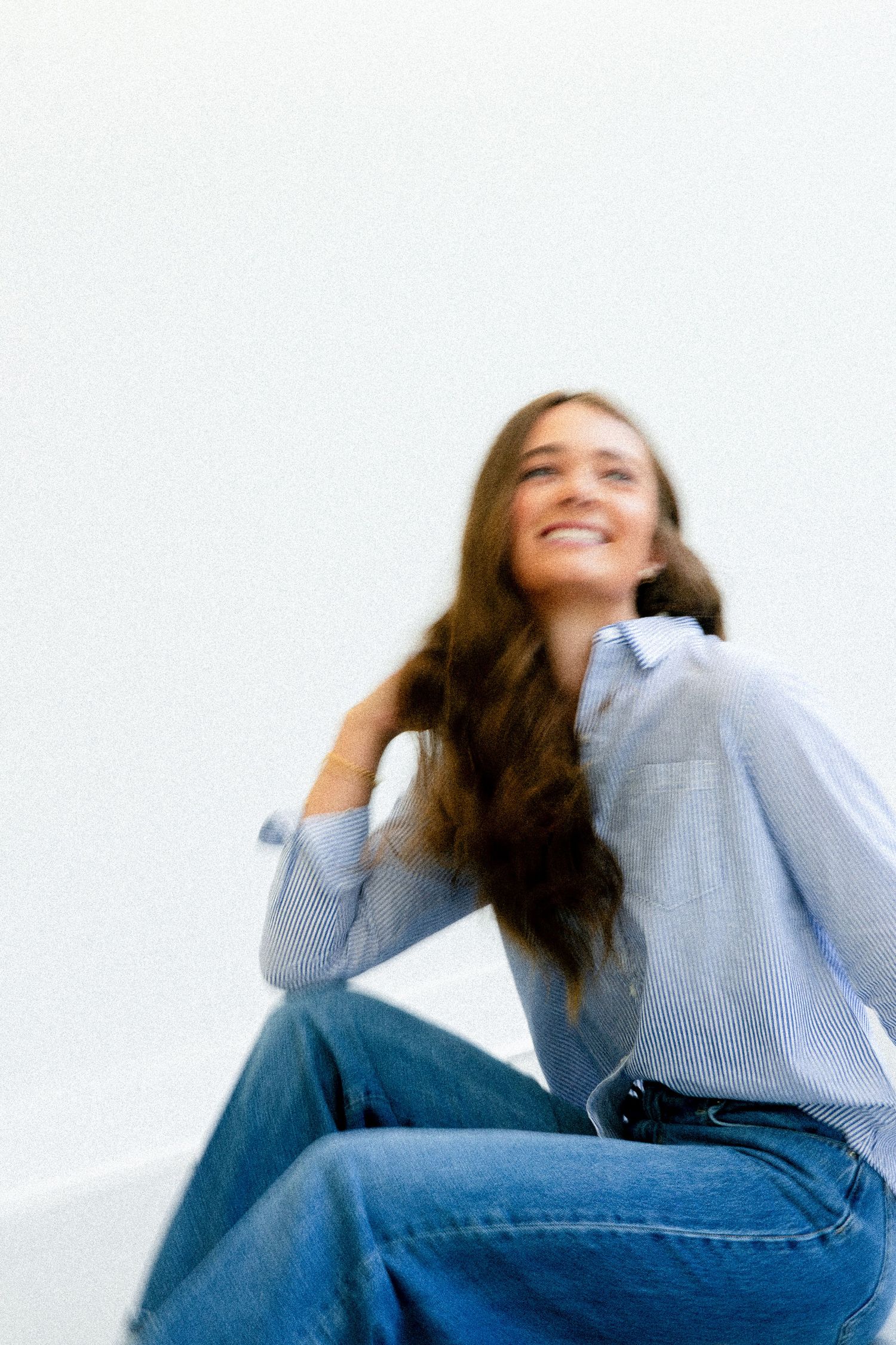 Smiling young woman with long brown hair wearing a blue striped shirt and wide-leg jeans, sitting against a white background.