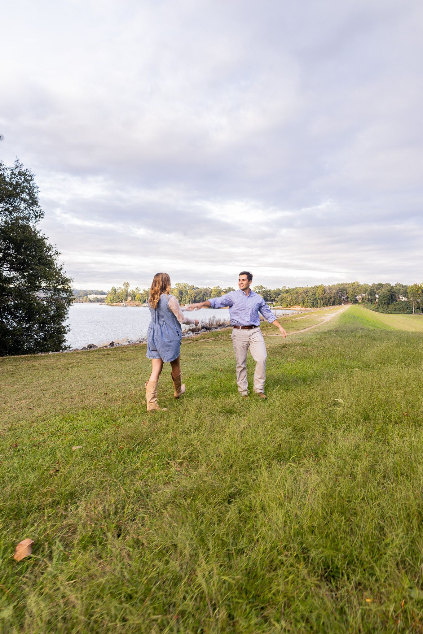 Couple holding hands and dancing on a grassy hillside near a lake under a cloudy sky.