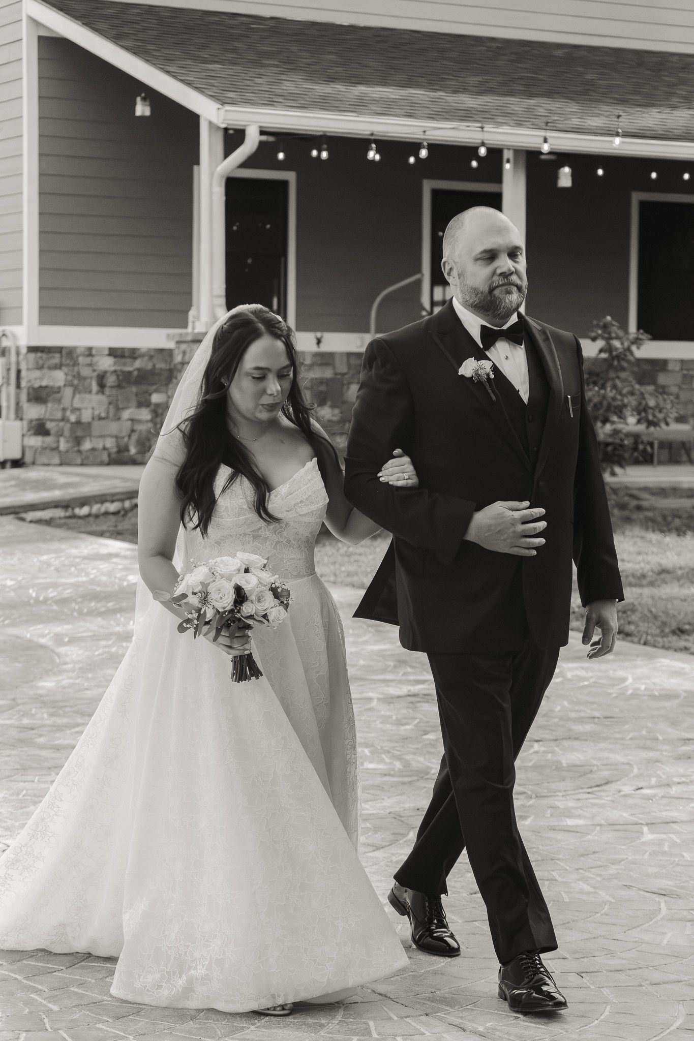 A bride and groom walk arm in arm in black and white photo outside a rustic wedding venue.