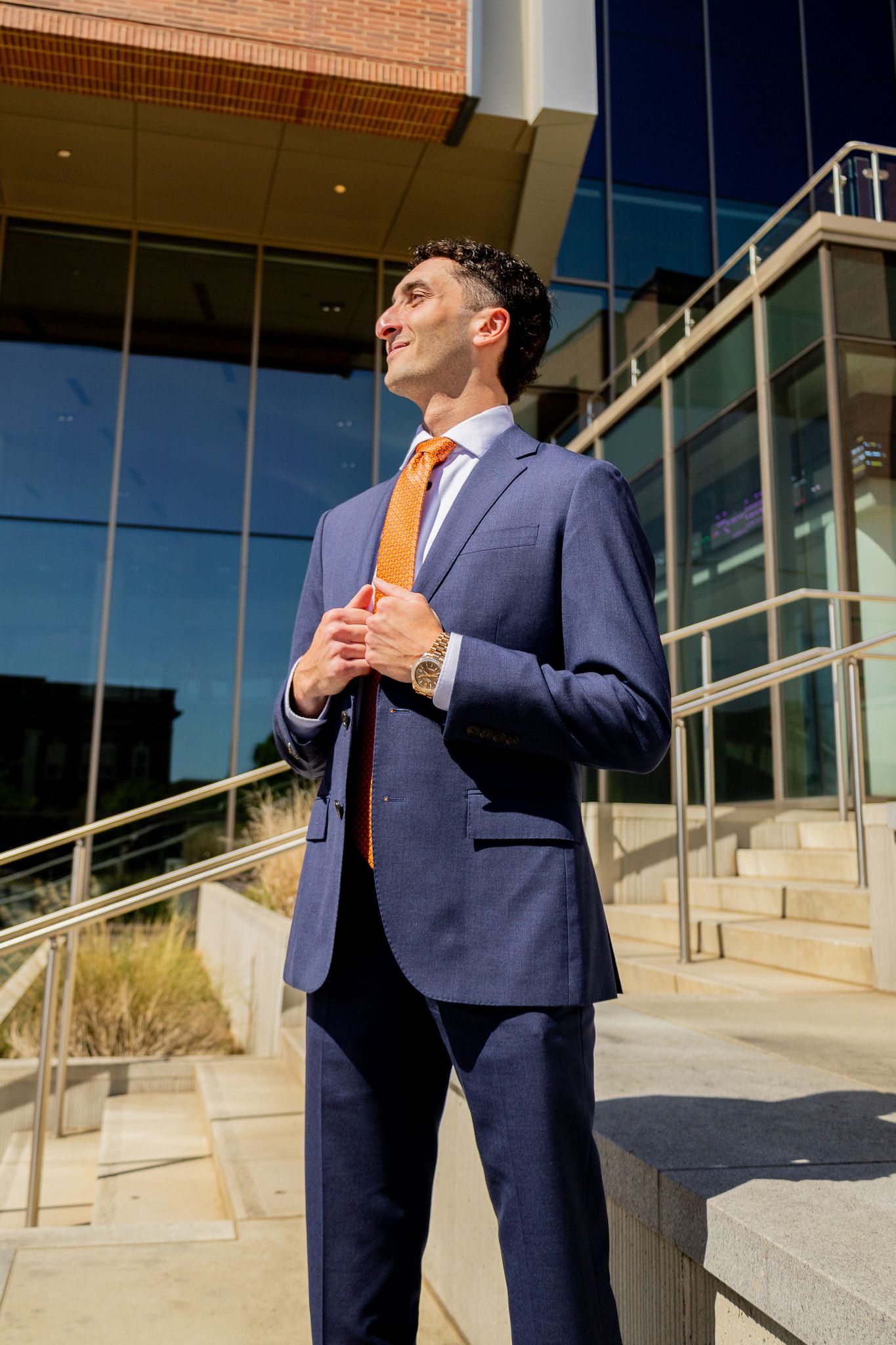 Professional man in navy blue suit and orange tie standing confidently outside a modern glass office building.