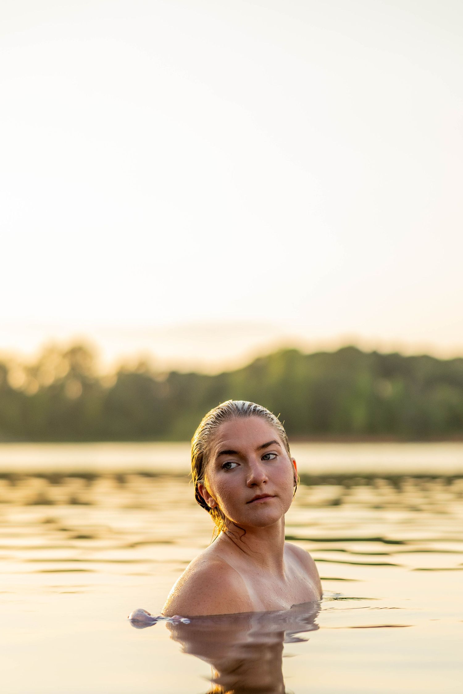 Woman swimming in a calm lake at golden hour sunset with lush green trees reflecting on the water.