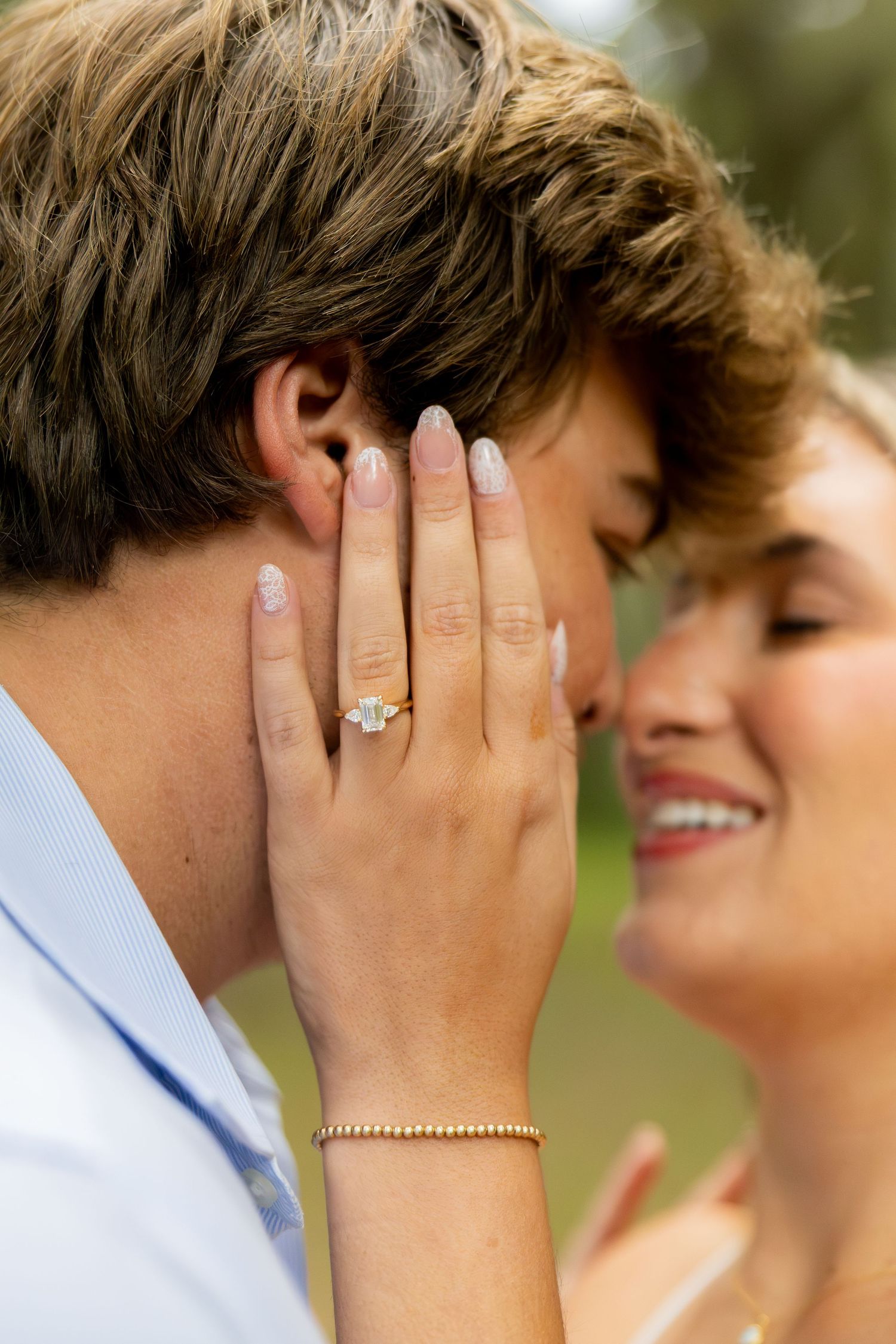 Close-up of a smiling couple embracing as a woman's hand with an engagement ring and gold bracelet rests on a man's cheek outdoors.