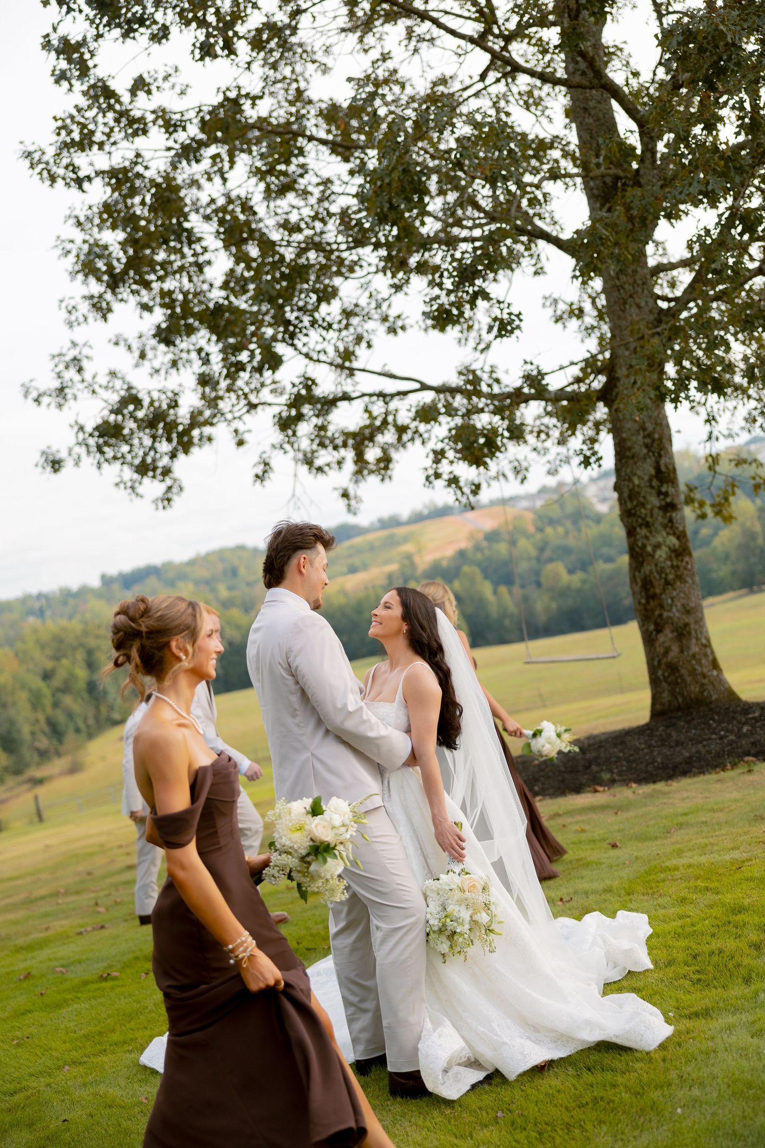Bride in white gown and veil stands with groom and bridesmaids in brown dresses on lush green lawn near large tree.