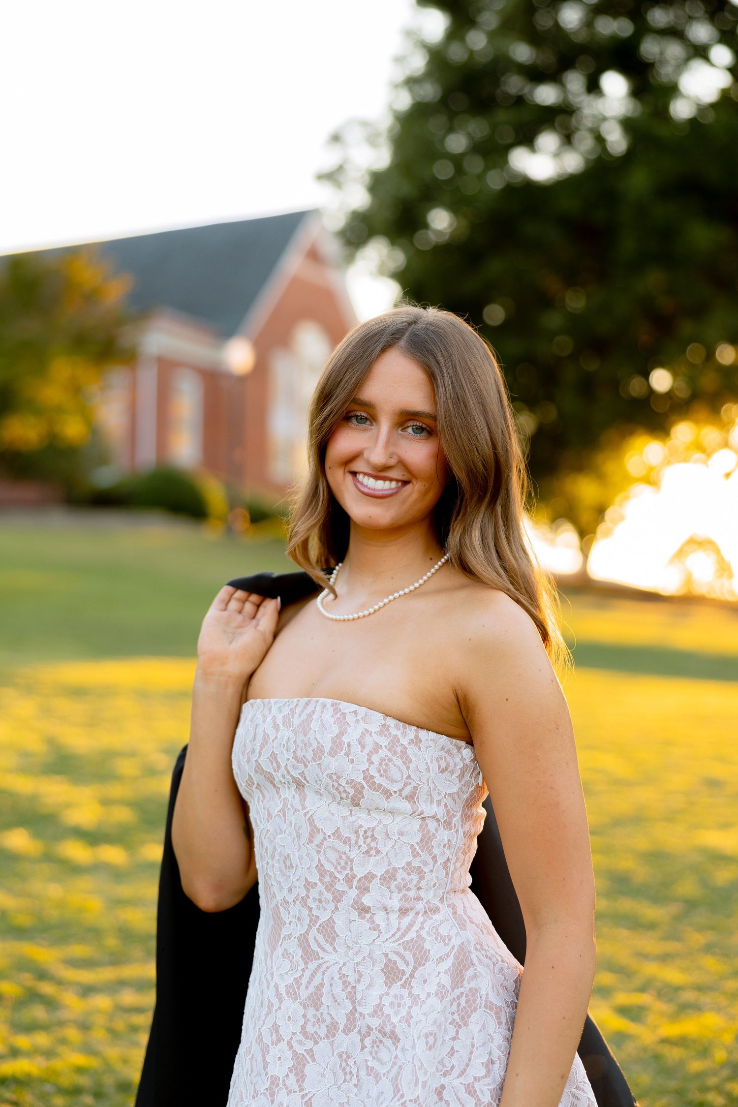 Young woman in white lace strapless dress and pearl necklace holding black gown outdoors at golden hour.
