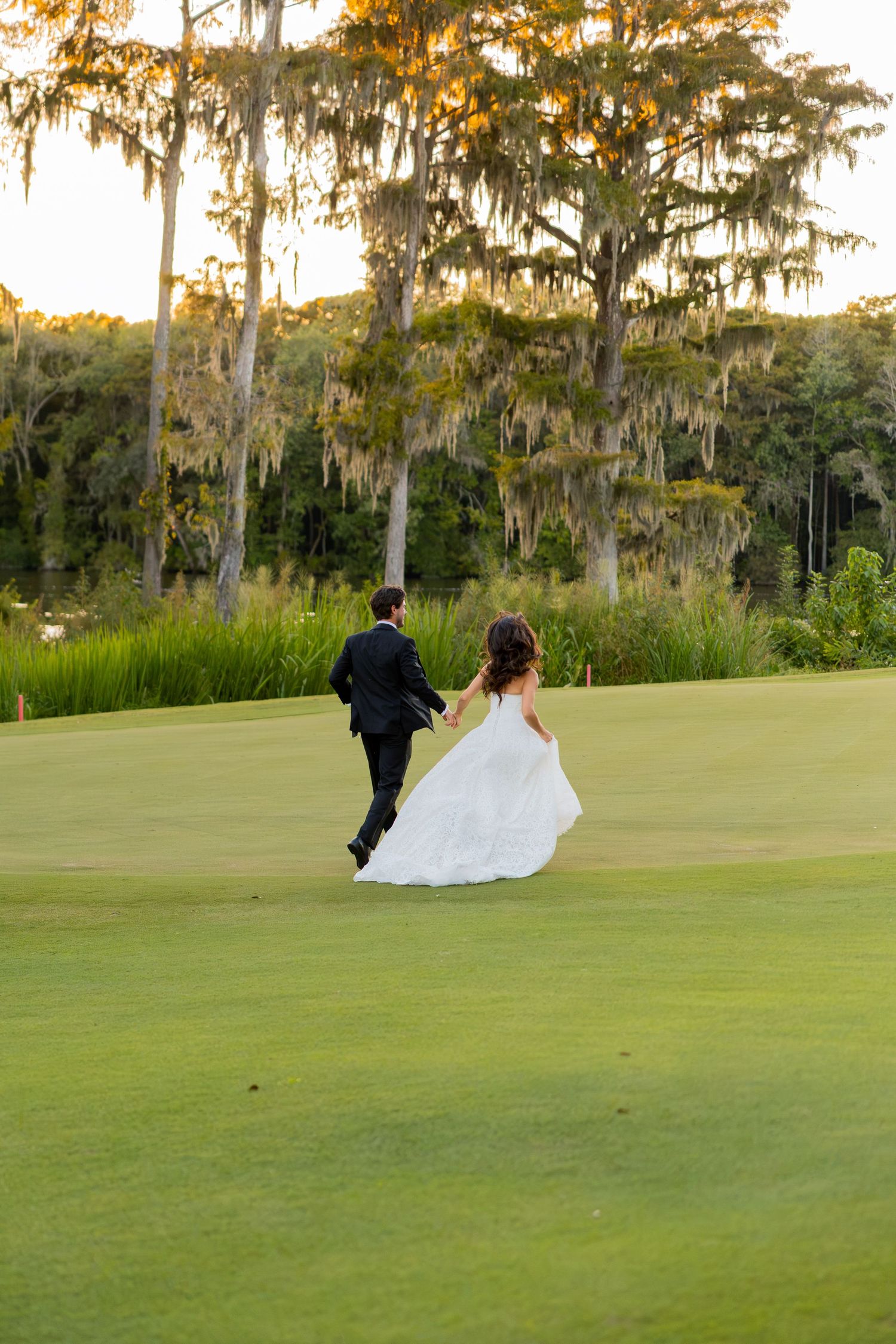 Bride and groom walking hand in hand across a lush green golf course at sunset, surrounded by moss-draped trees.