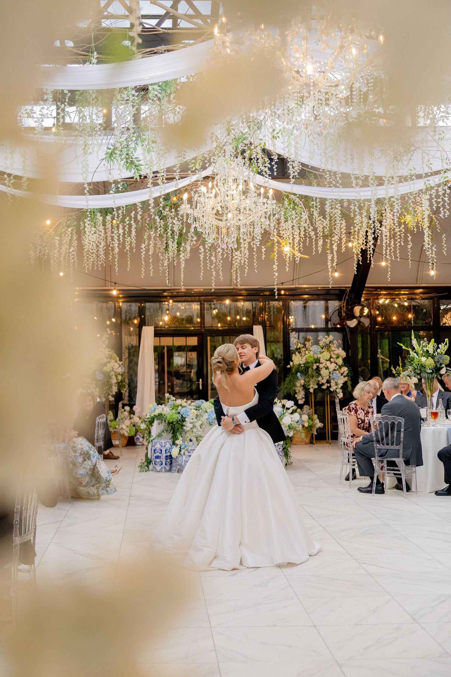 Bride and groom share first dance under floral chandeliers and draped lights in an elegant wedding reception venue.
