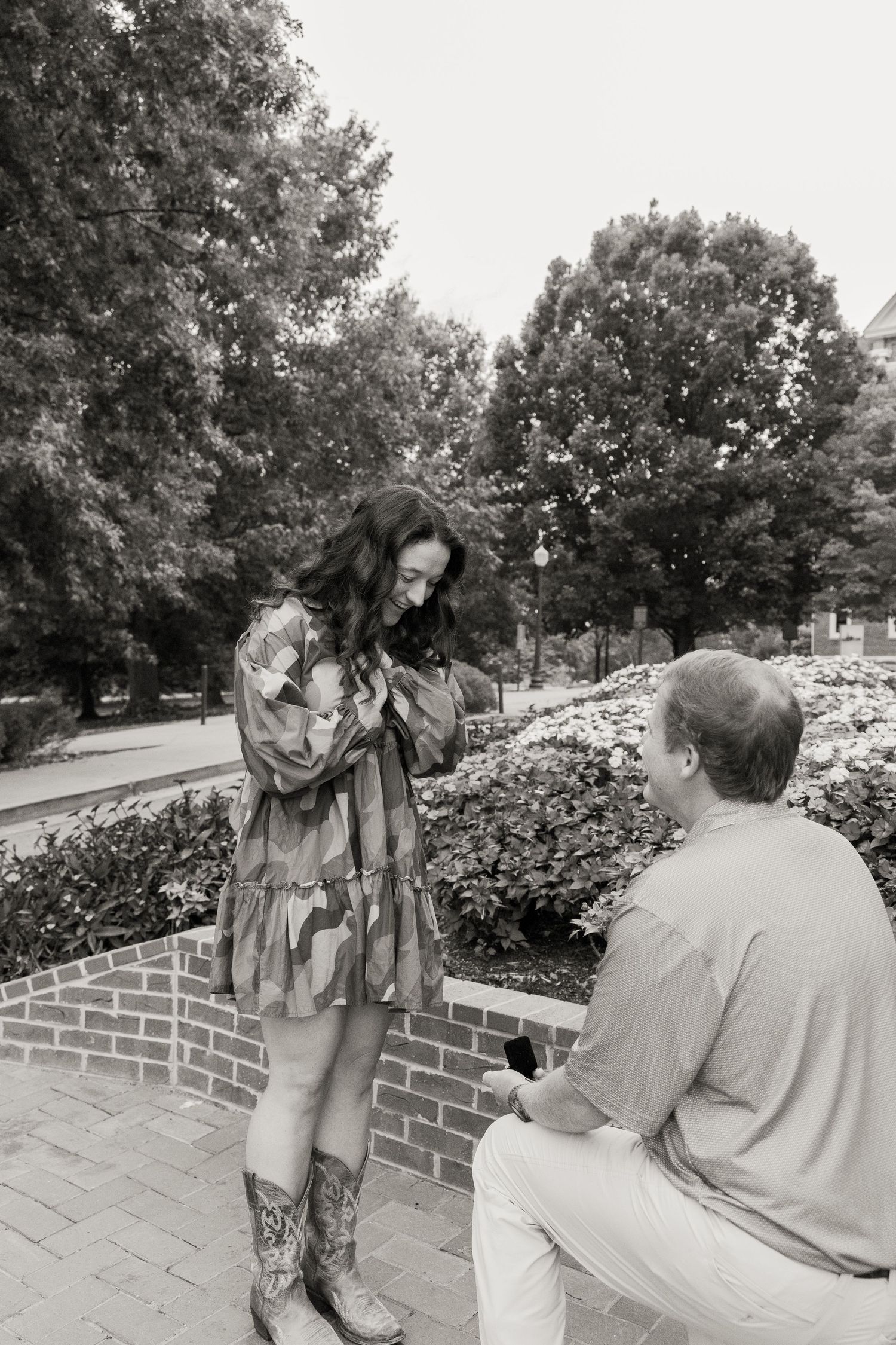 Man proposing on one knee to woman in floral dress outdoors, black and white photo.