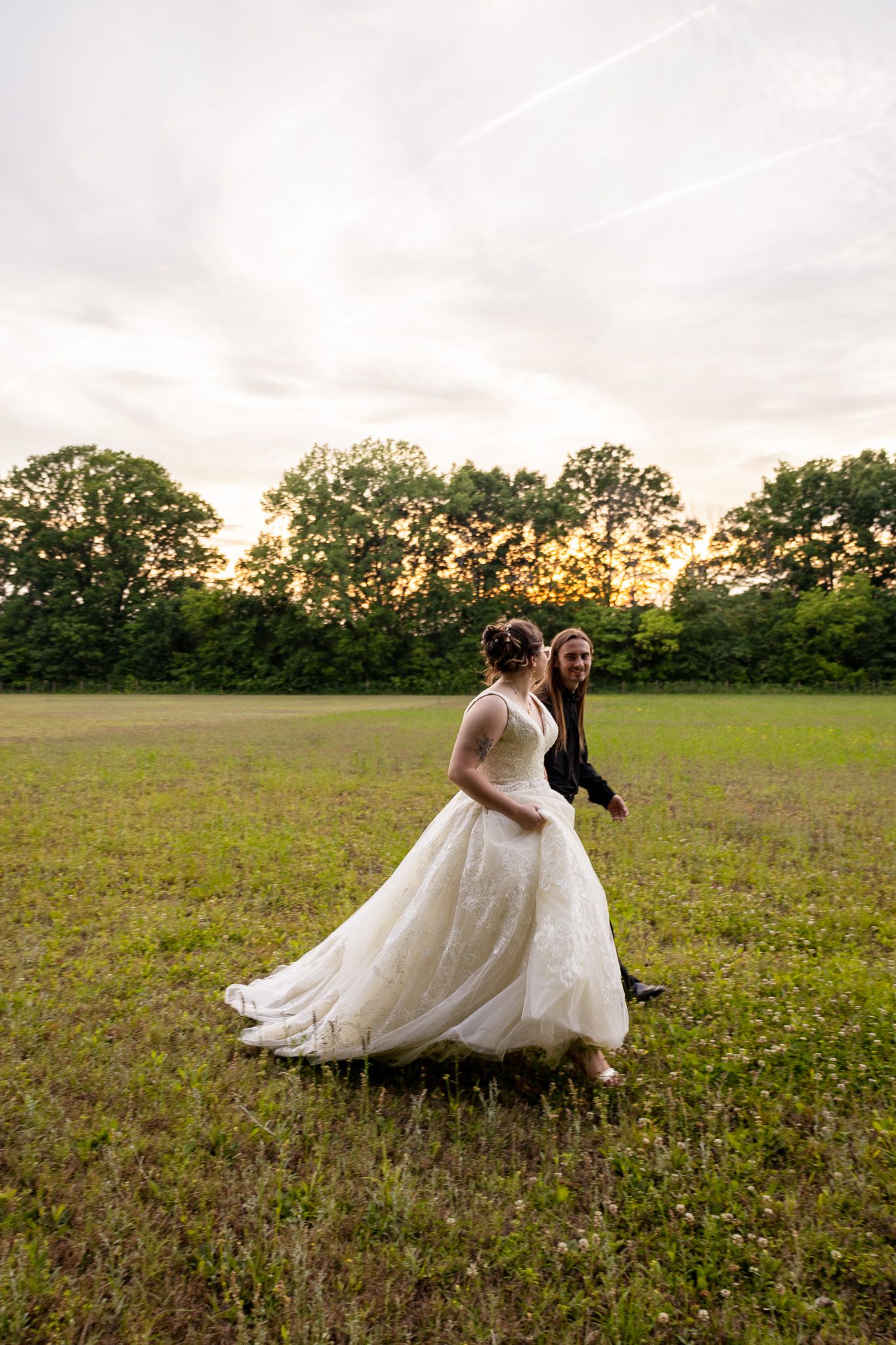 Two brides walk through a green field at sunset, one in a white ball gown and one in a black jacket, smiling joyfully.