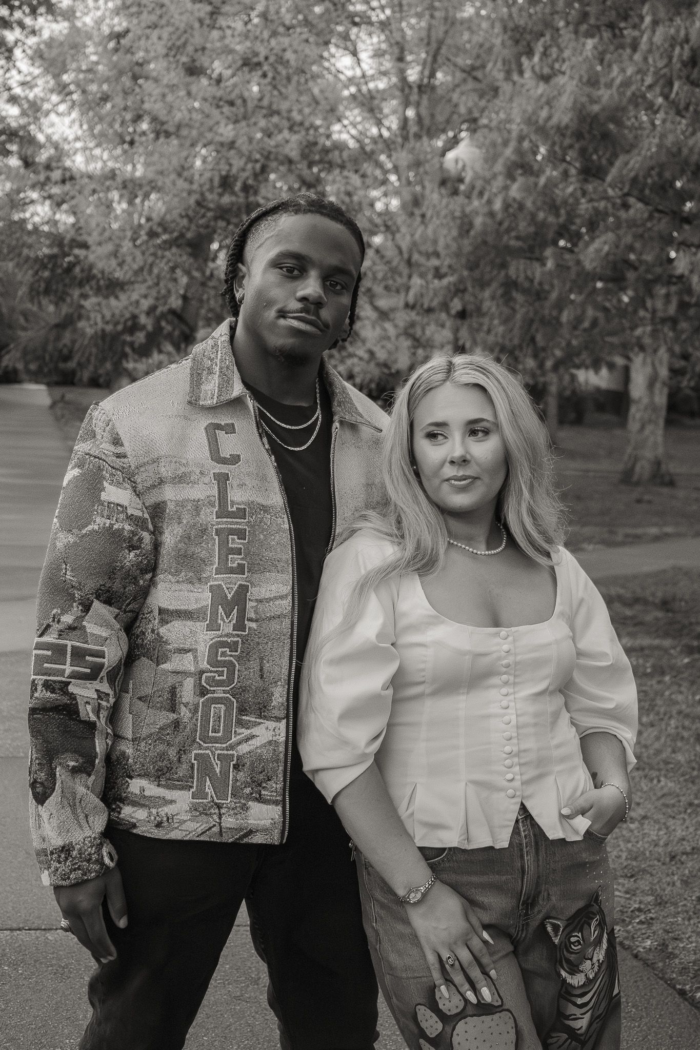 Black and white photo of a couple posing outdoors, man wearing a Clemson jacket, woman in white top and jeans.