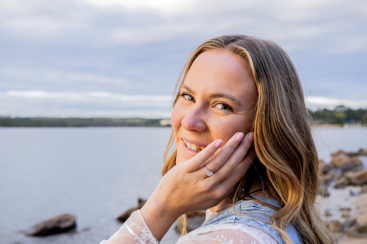 Smiling blonde woman touching her face near a serene waterfront, wearing a white lace top on a cloudy day.