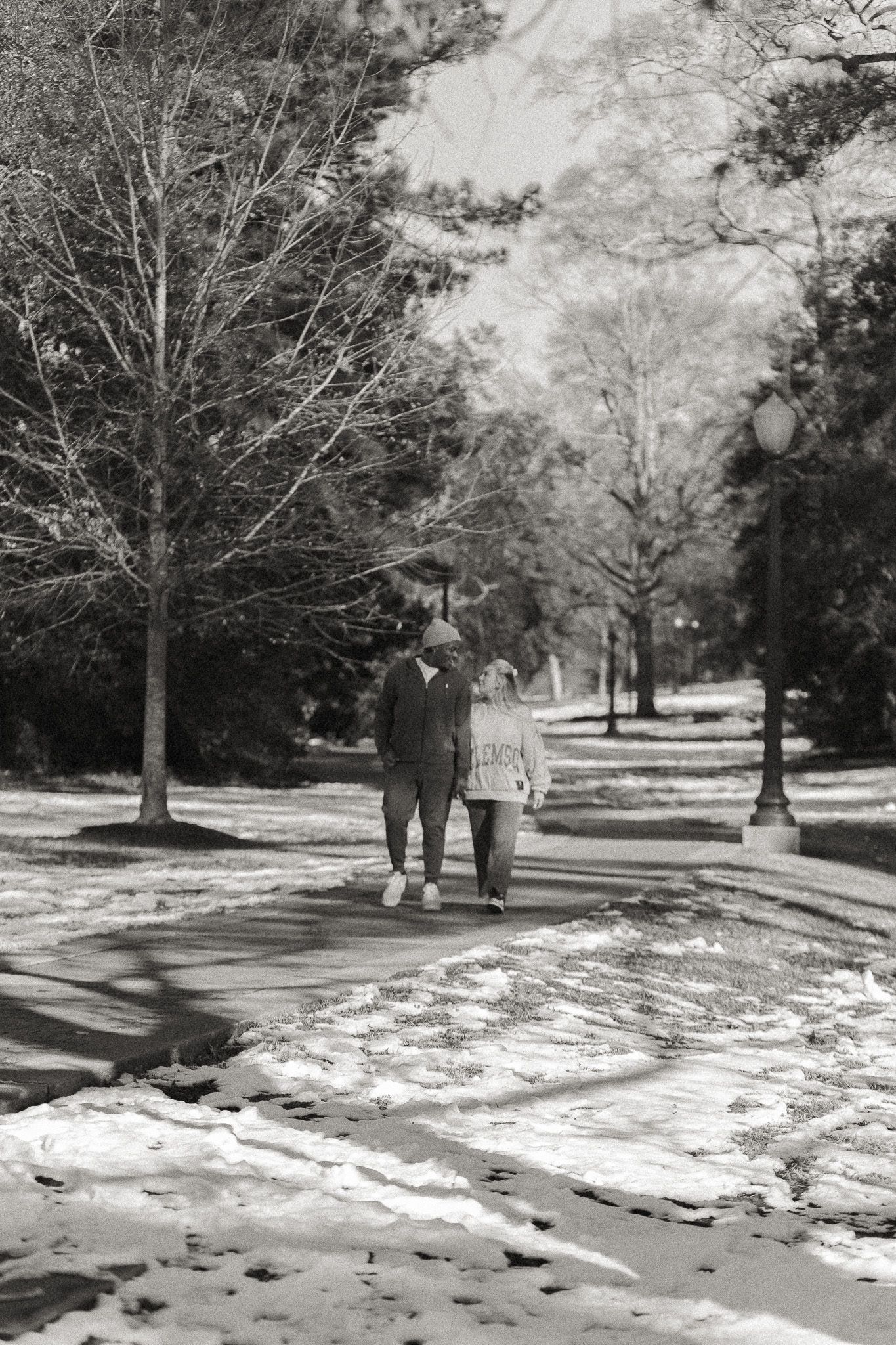 Couple walking hand in hand on a snowy park path lined with bare trees in a black and white winter scene.