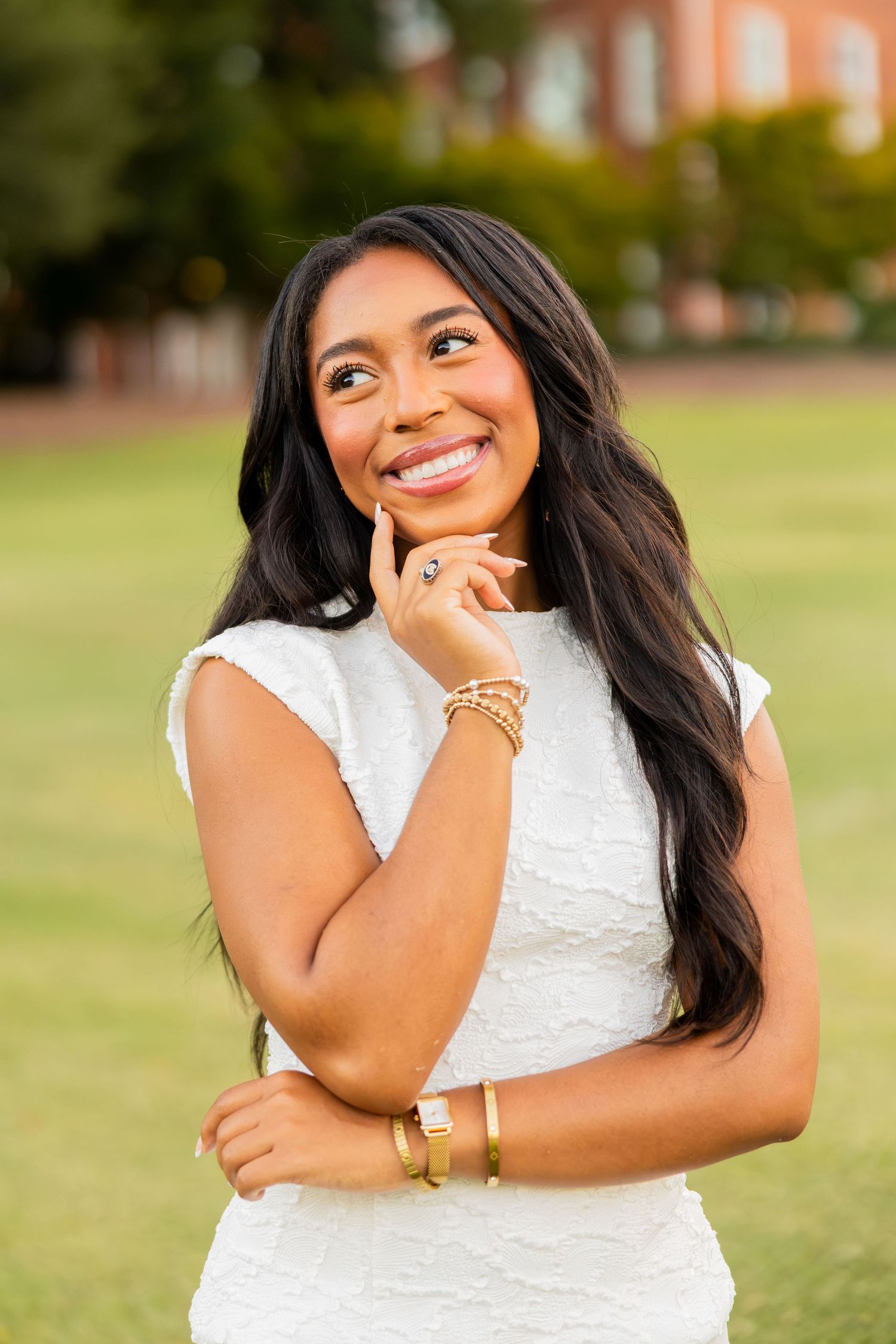 Smiling young woman in white dress with gold jewelry, posing outdoors on green lawn near brick building.