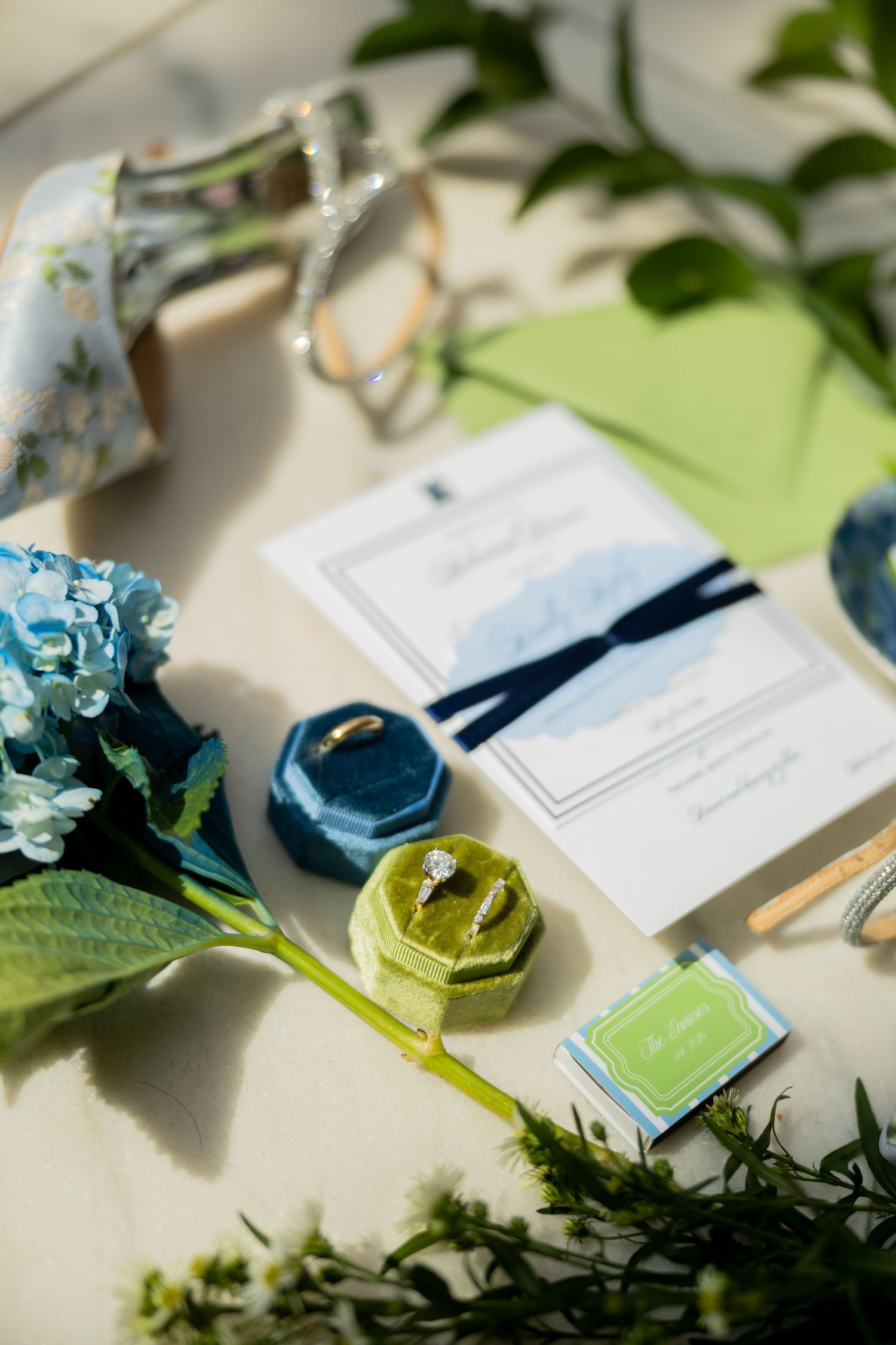 Wedding flat lay with diamond ring in green velvet box, blue hydrangeas, invitation suite, and floral heels on cream background.