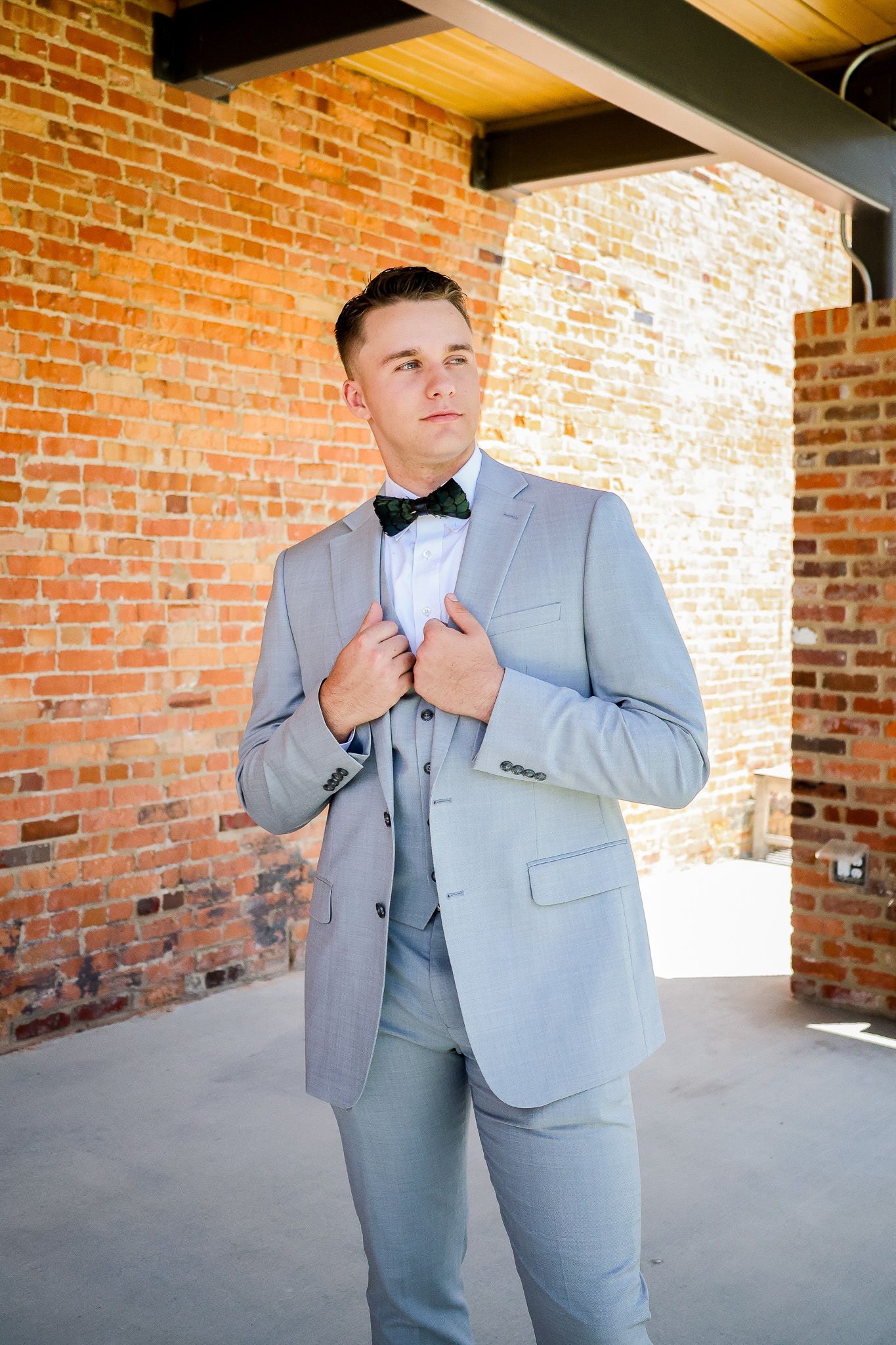 Young groom in light gray suit and black bow tie adjusts jacket lapels against rustic brick wall backdrop.