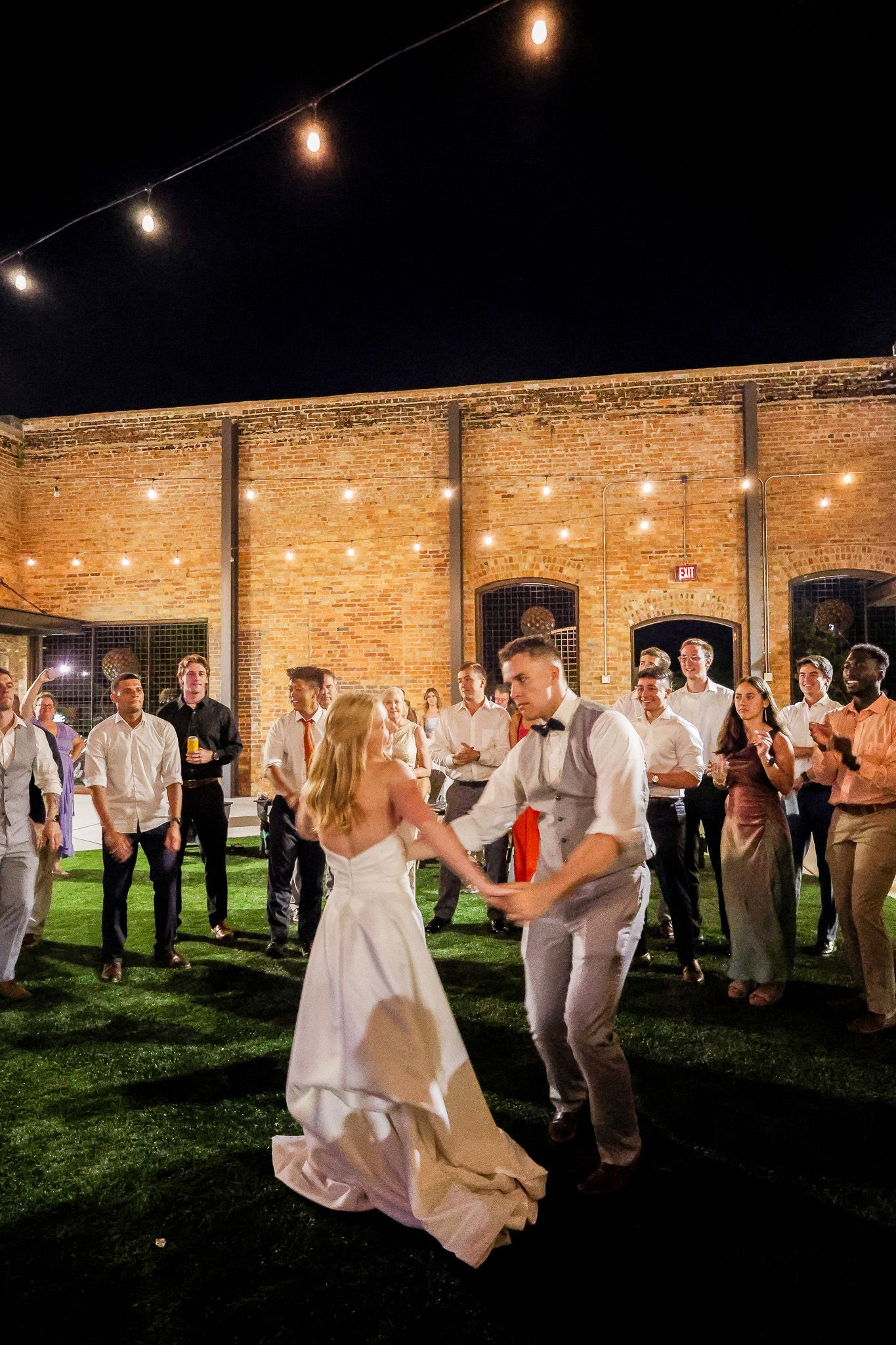 Bride and groom share first dance outdoors at night, surrounded by guests near a brick wall with string lights.