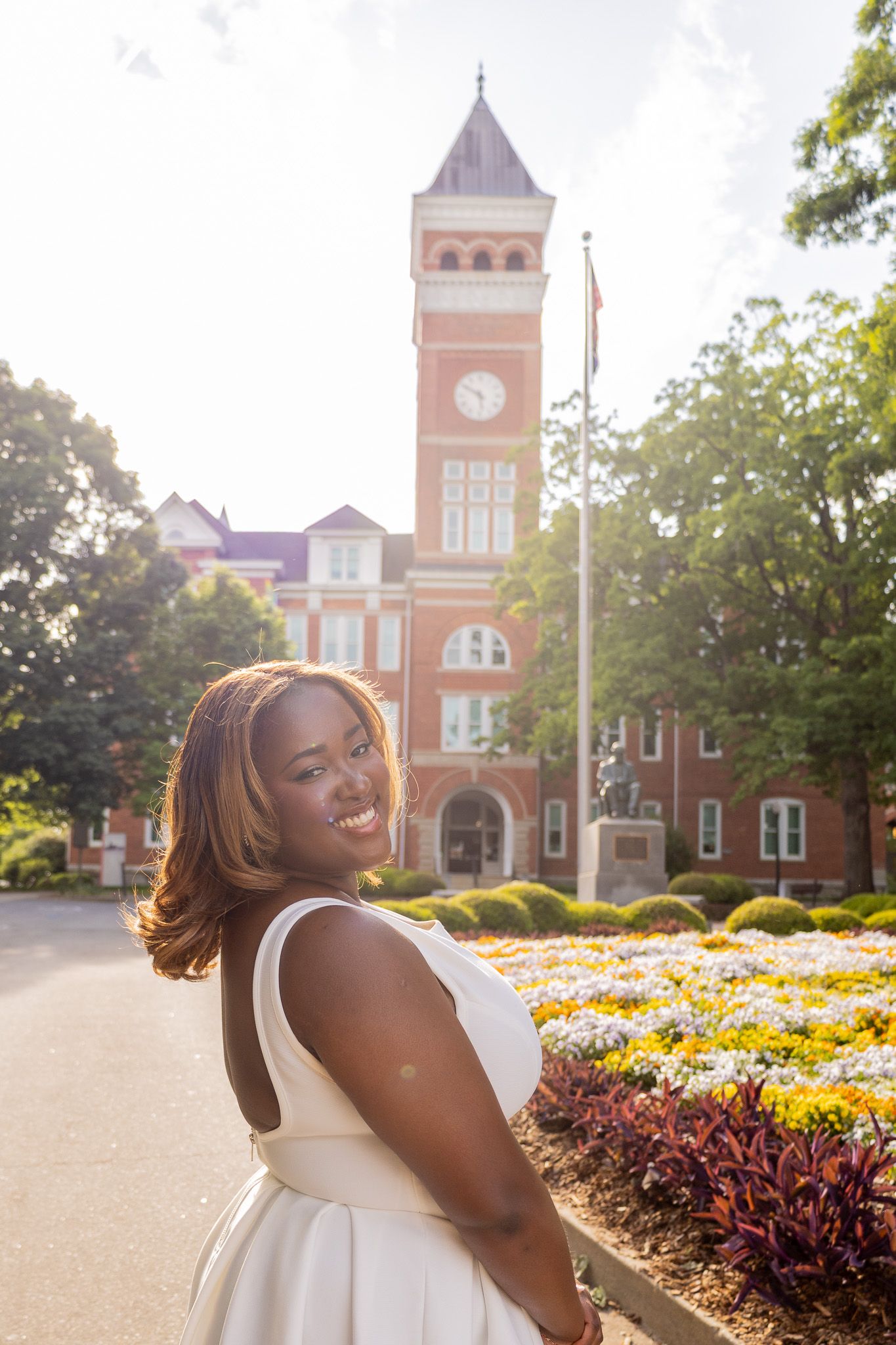 Smiling Black woman in white dress poses before a red brick clock tower building with colorful flowers.