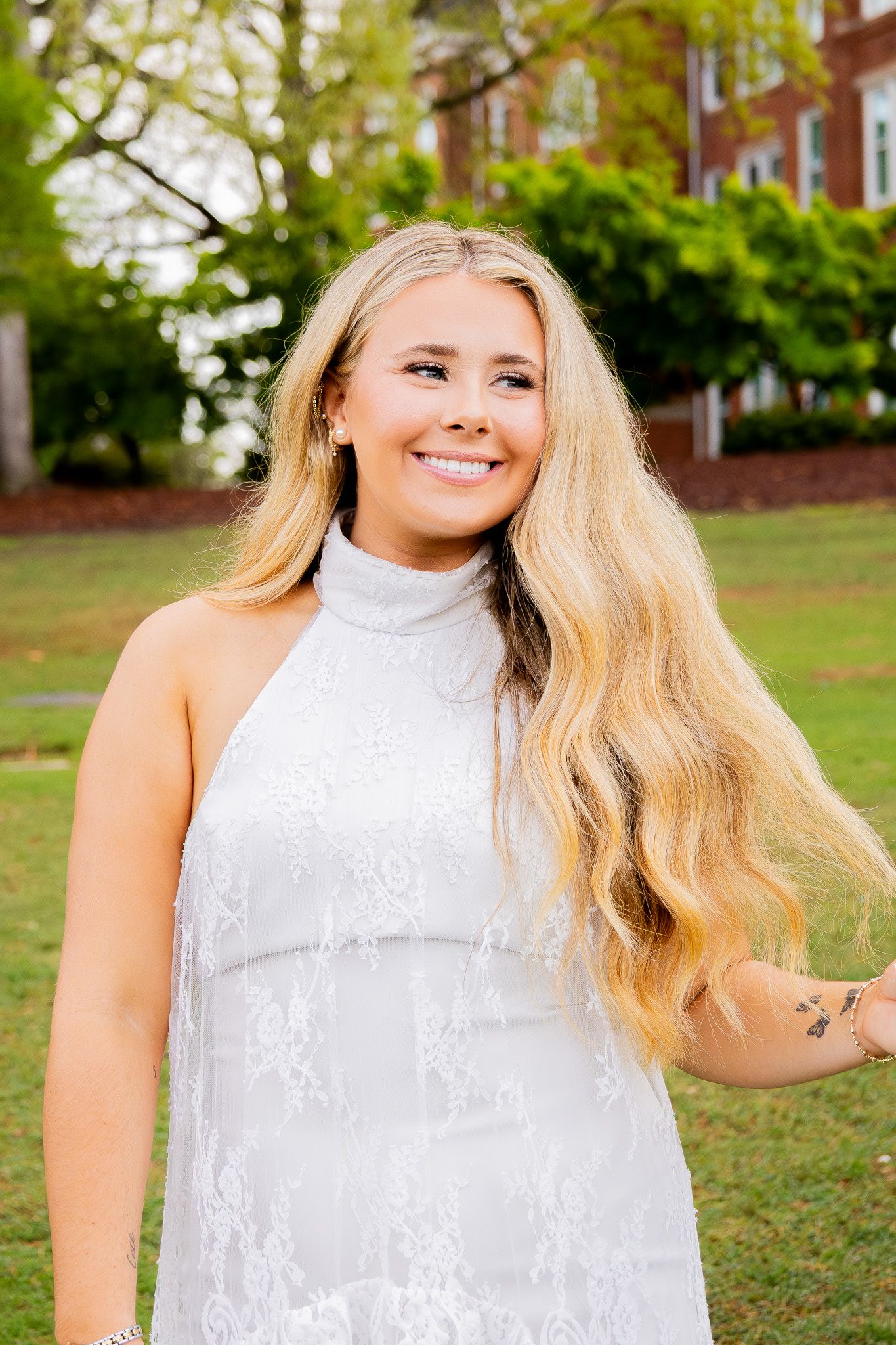 Young blonde woman in white lace halter dress smiling outdoors on green lawn with trees and brick building.