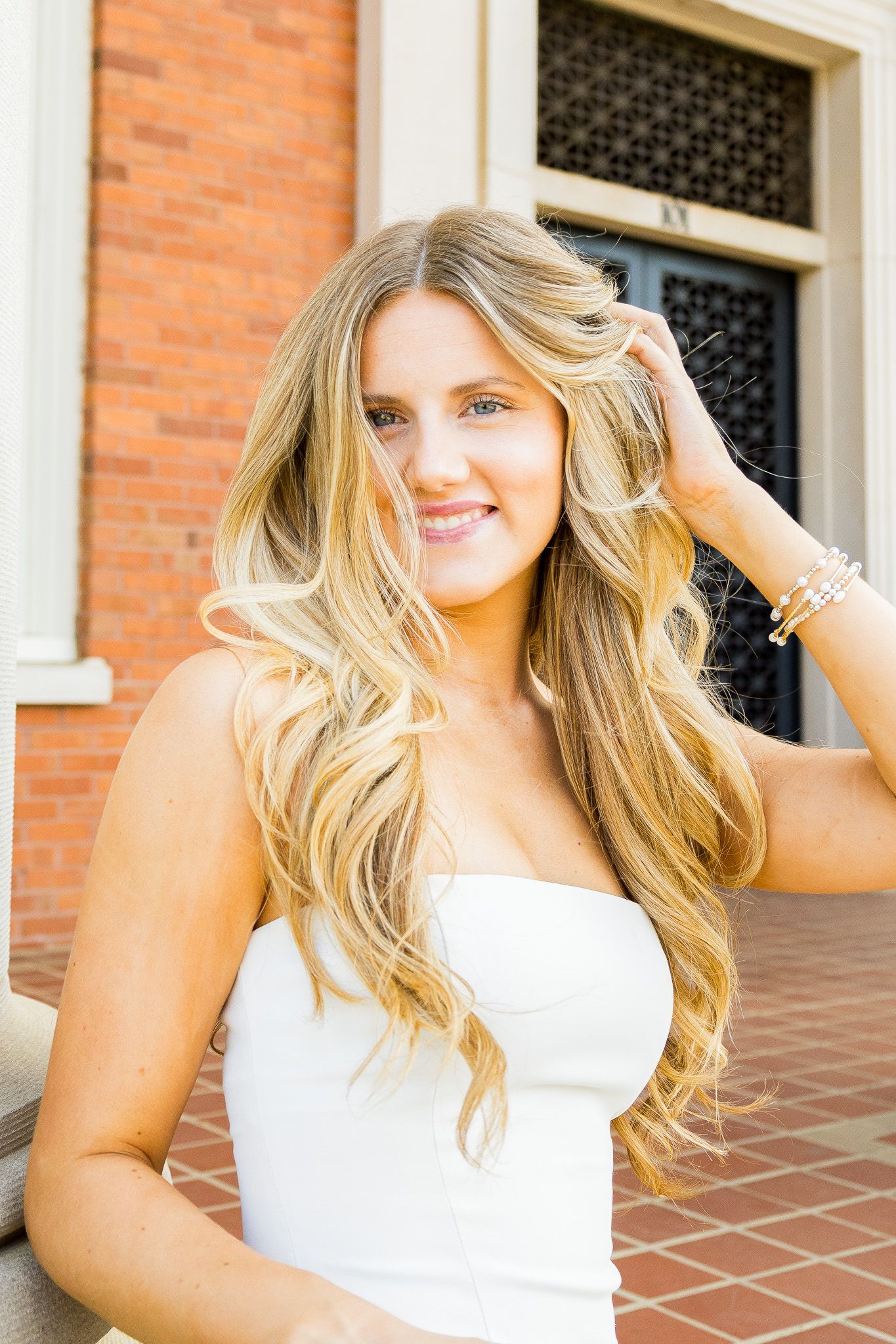 Young blonde woman in white strapless dress smiling outdoors near brick building, hand in hair.