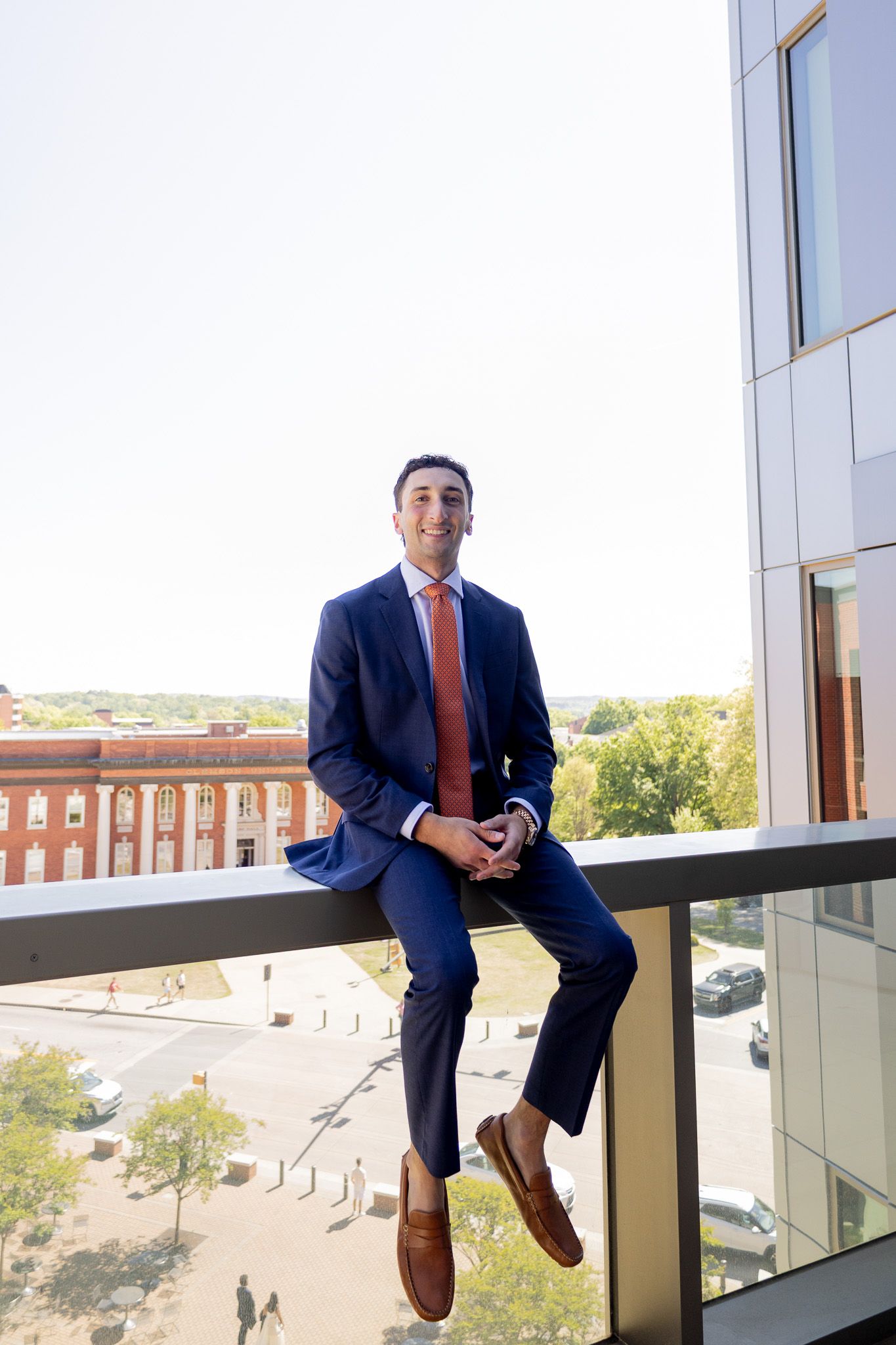 Young professional man in navy suit and orange tie sitting on building balcony railing, smiling confidently outdoors.
