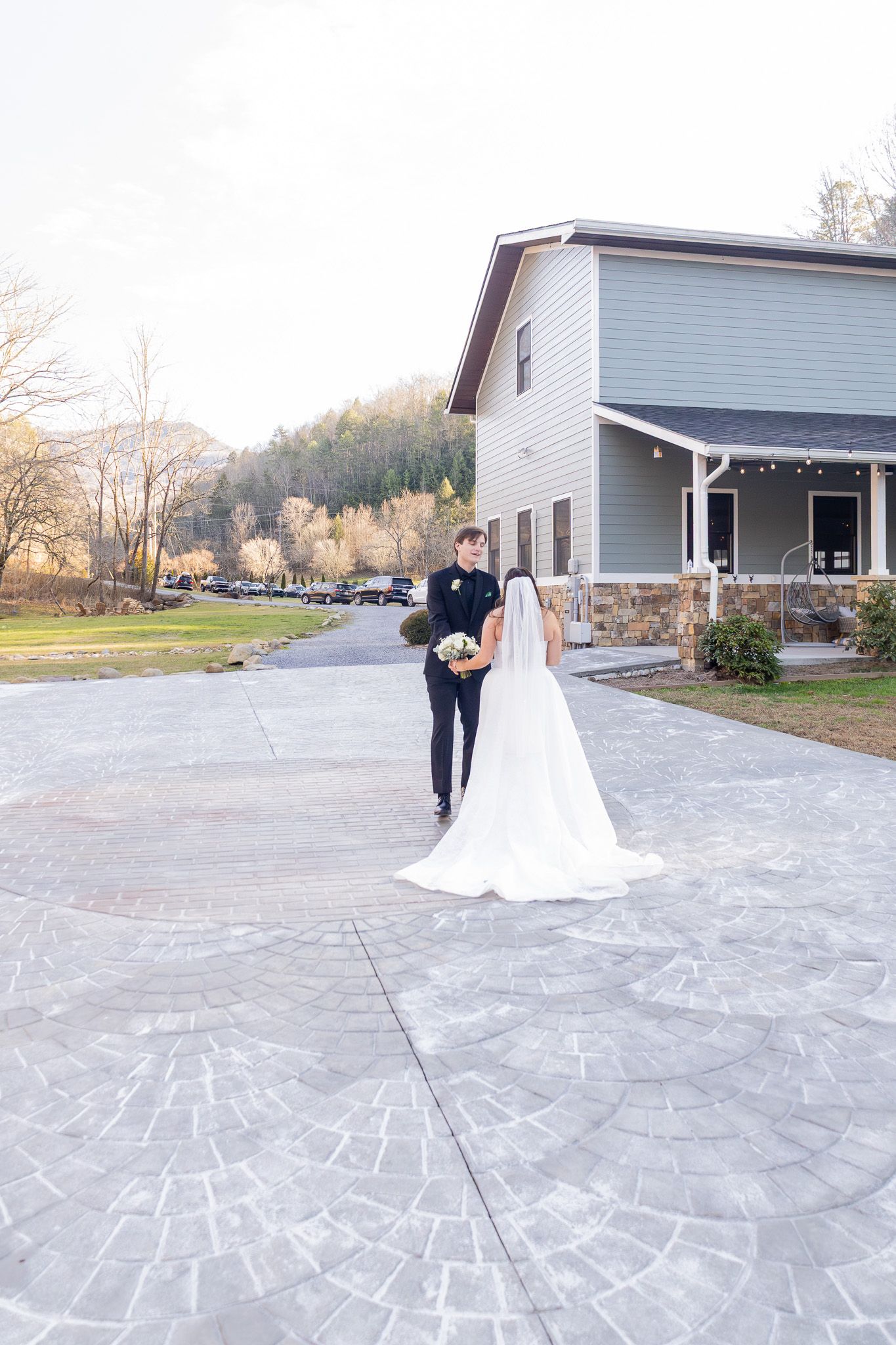 Bride in white gown and groom in black suit walk toward a venue building on a paved driveway surrounded by autumn trees.