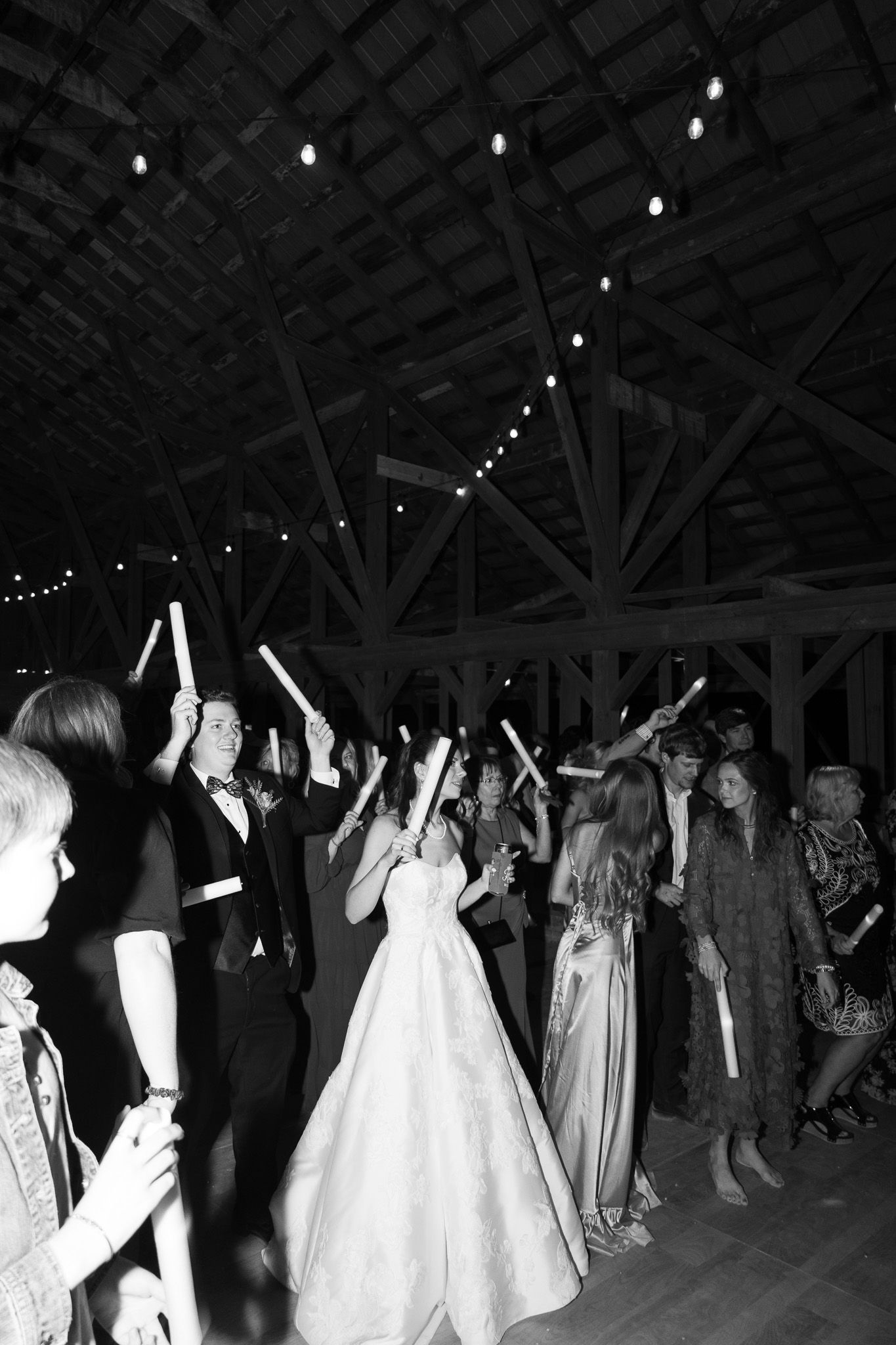 Bride and groom celebrate with guests waving glow sticks at an elegant barn wedding reception.