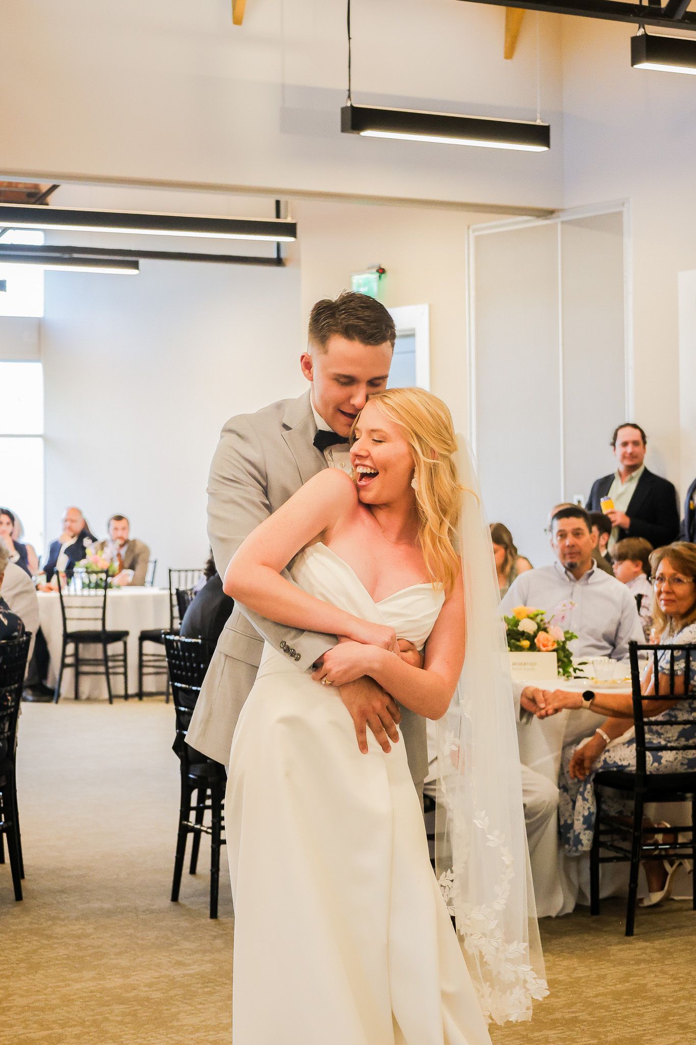 Joyful bride in white gown laughs as groom in gray suit embraces her during wedding reception.