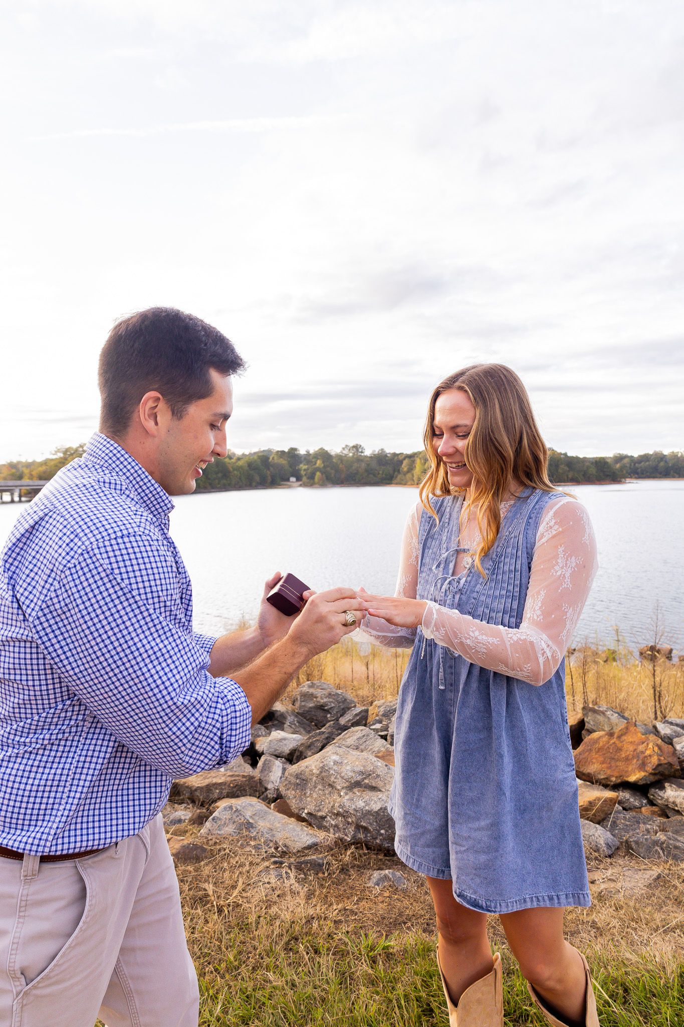 Man in blue shirt presents engagement ring to smiling woman in denim dress by a scenic lakeside.