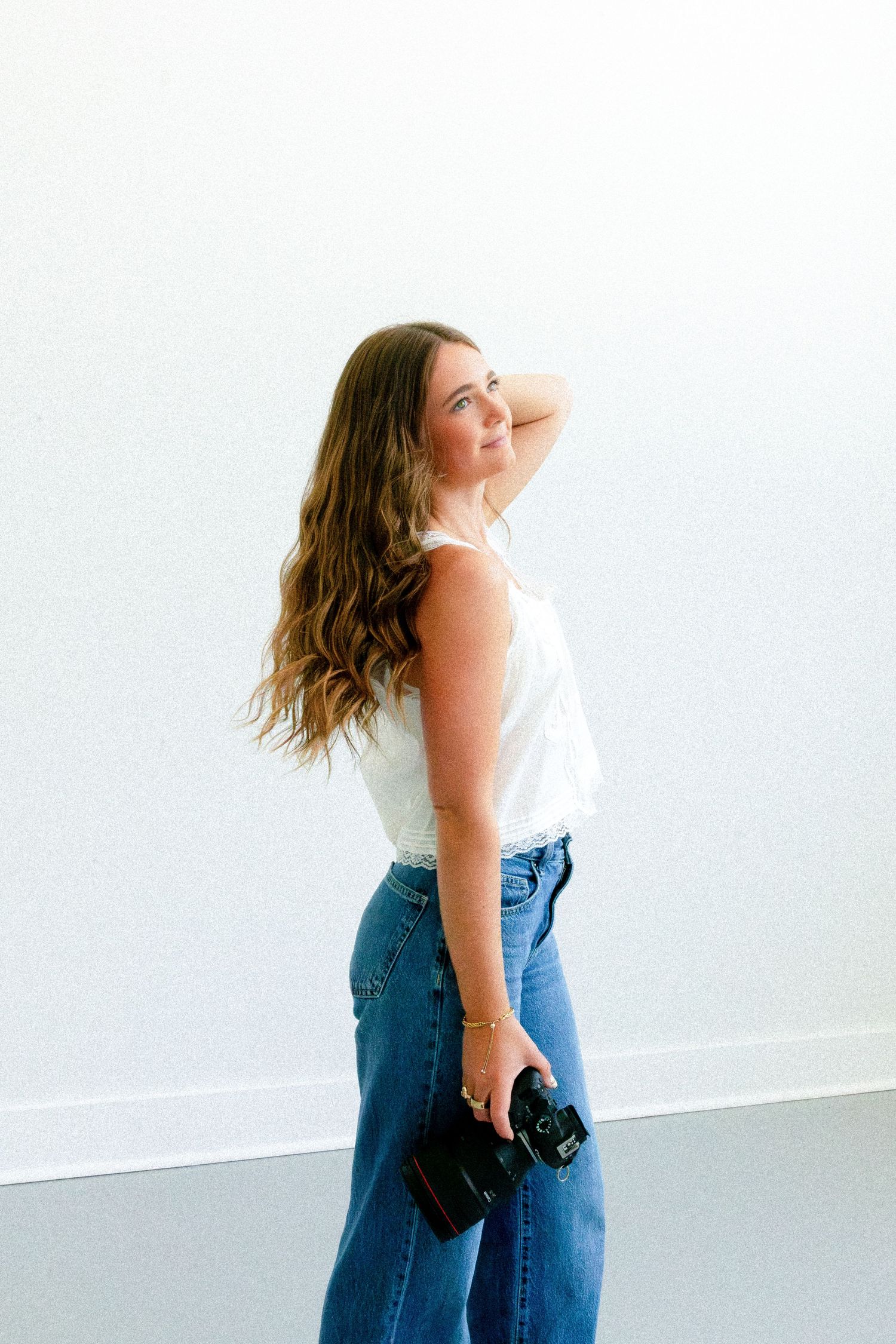 Young woman in white tank top and blue jeans holding a camera, posing against a bright white wall.