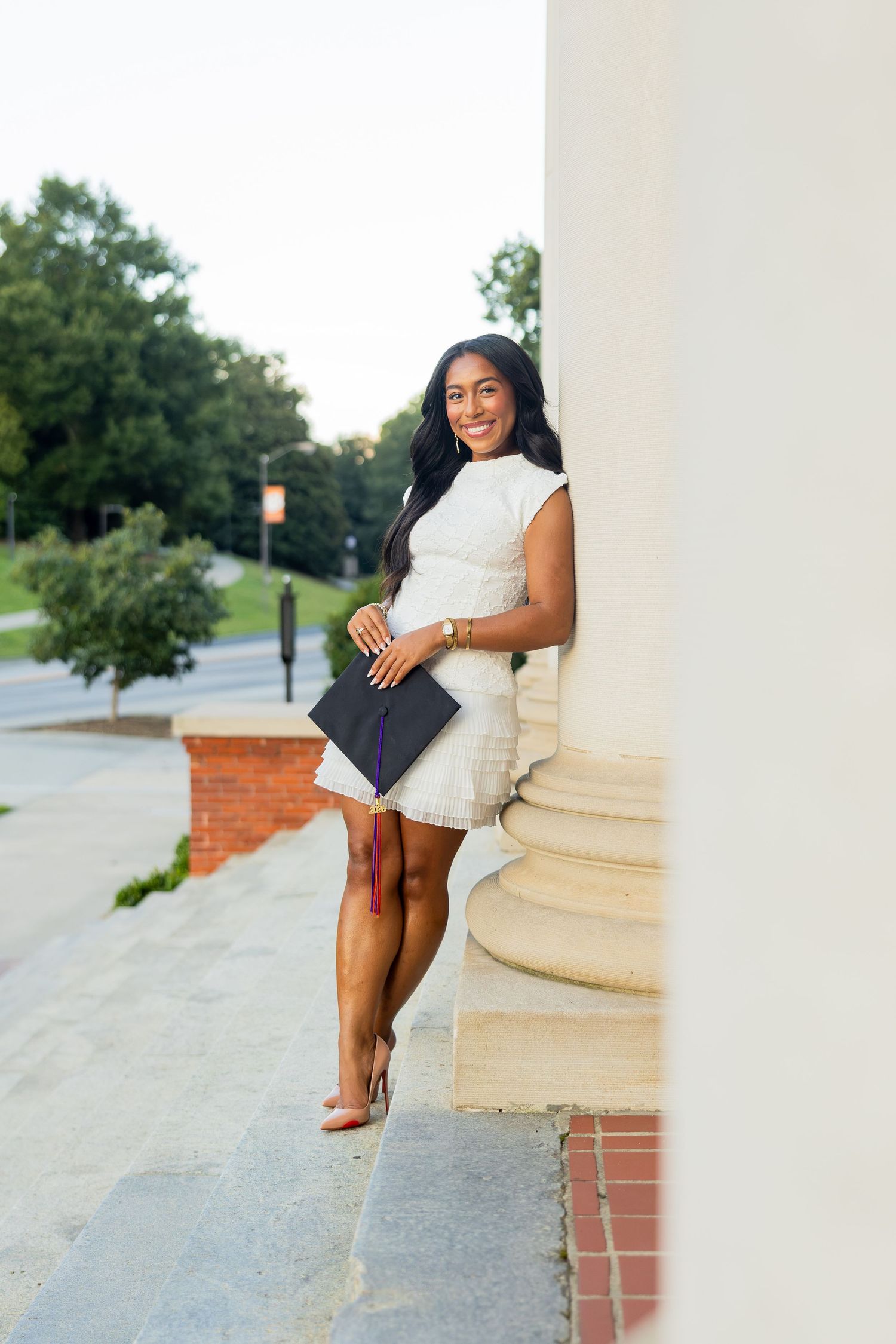 Smiling young woman in white dress holding graduation cap, leaning against column outdoors on campus.