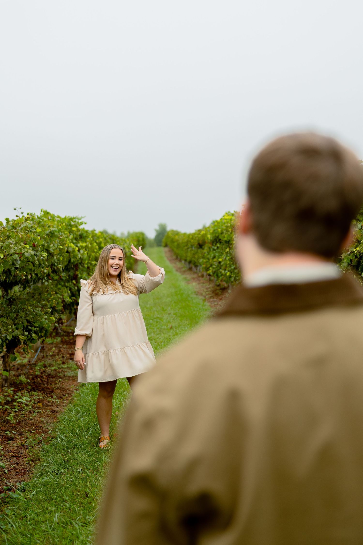 A smiling woman in a white dress waves to a man in a vineyard on an overcast day.